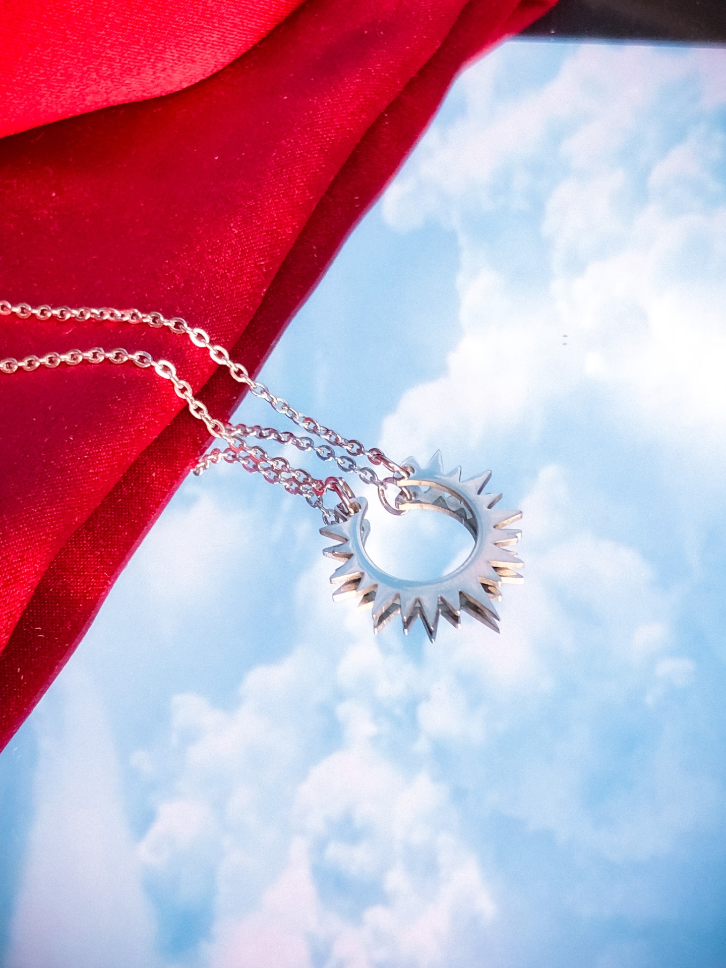 Colar prateado com pendente circular pontiagudo sobre fundo de céu com nuvens e tecido vermelho