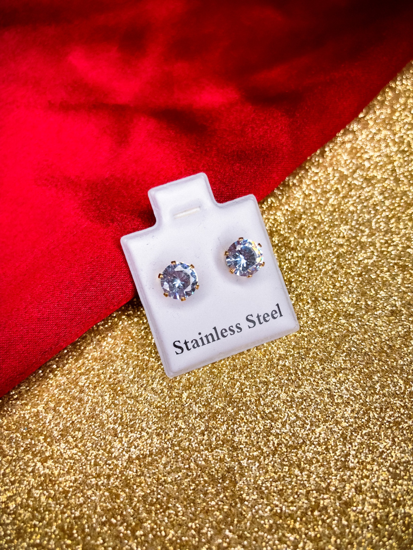 Par de brincos com pedras transparentes em cartão branco sobre fundo dourado e vermelho