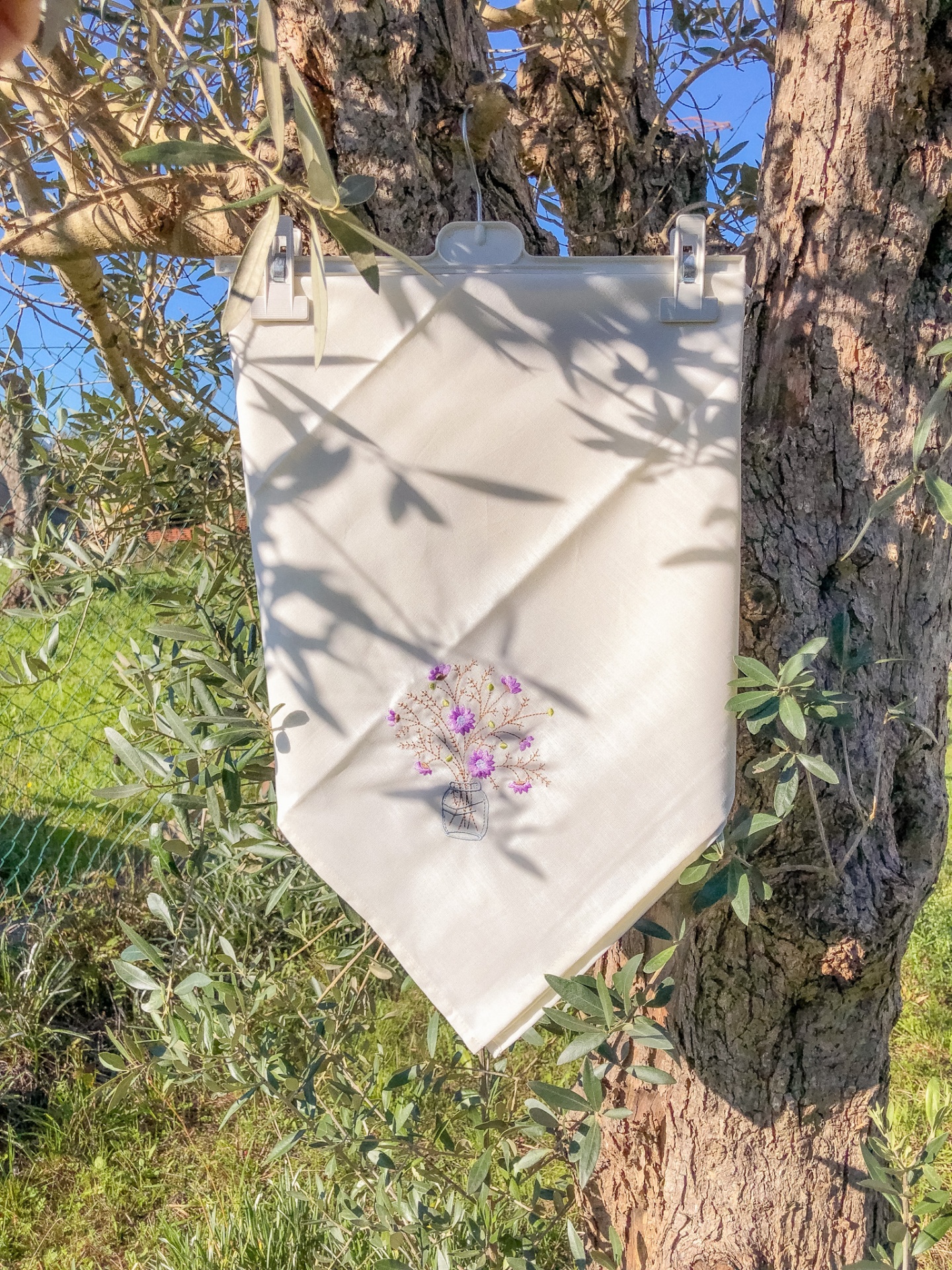 Pano branco com bordado de flores roxas pendurado numa árvore