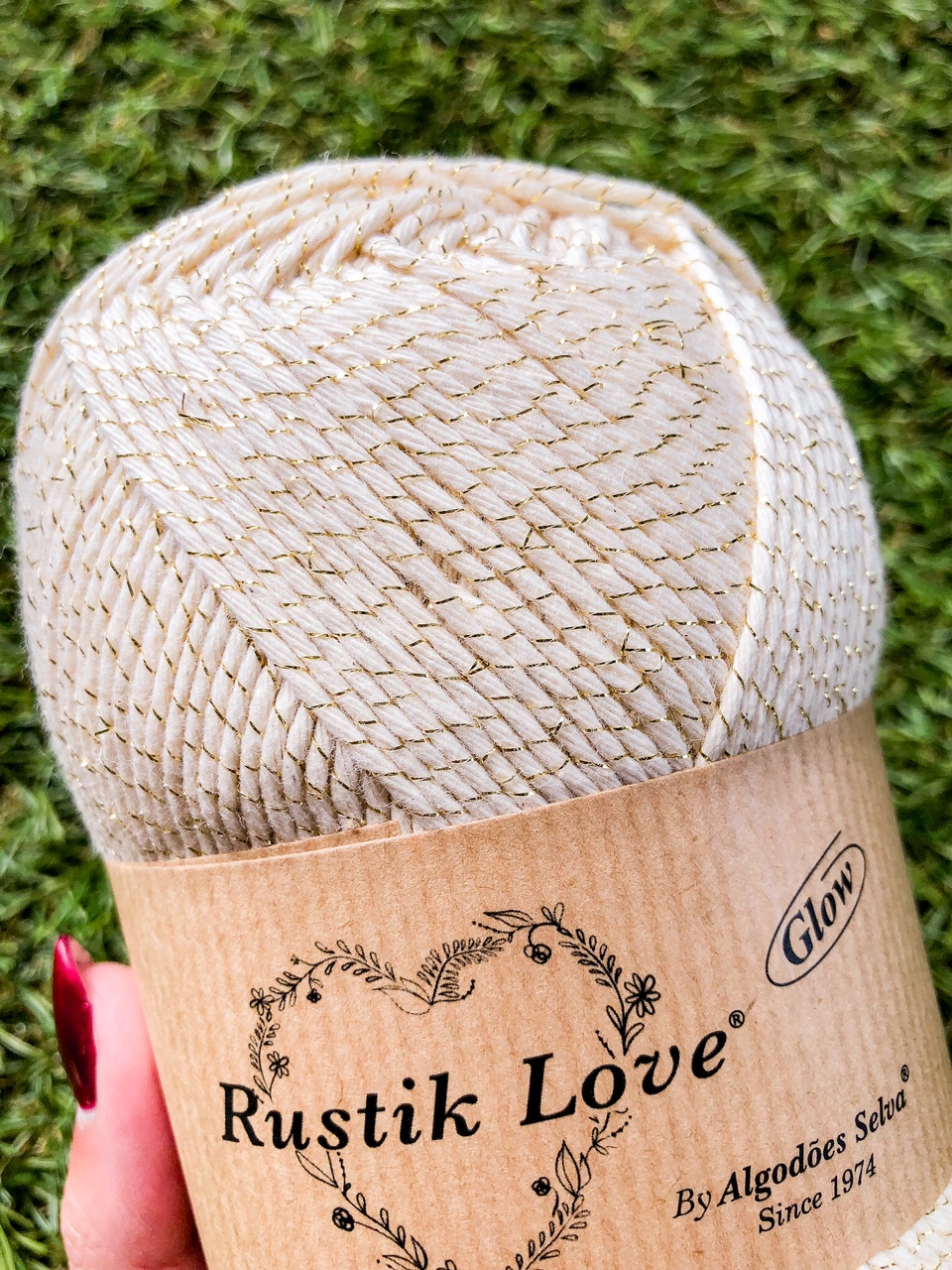 Novelo de fio Rustik Love cor creme com brilho dourado e etiqueta de papel kraft em fundo de relva verde.
