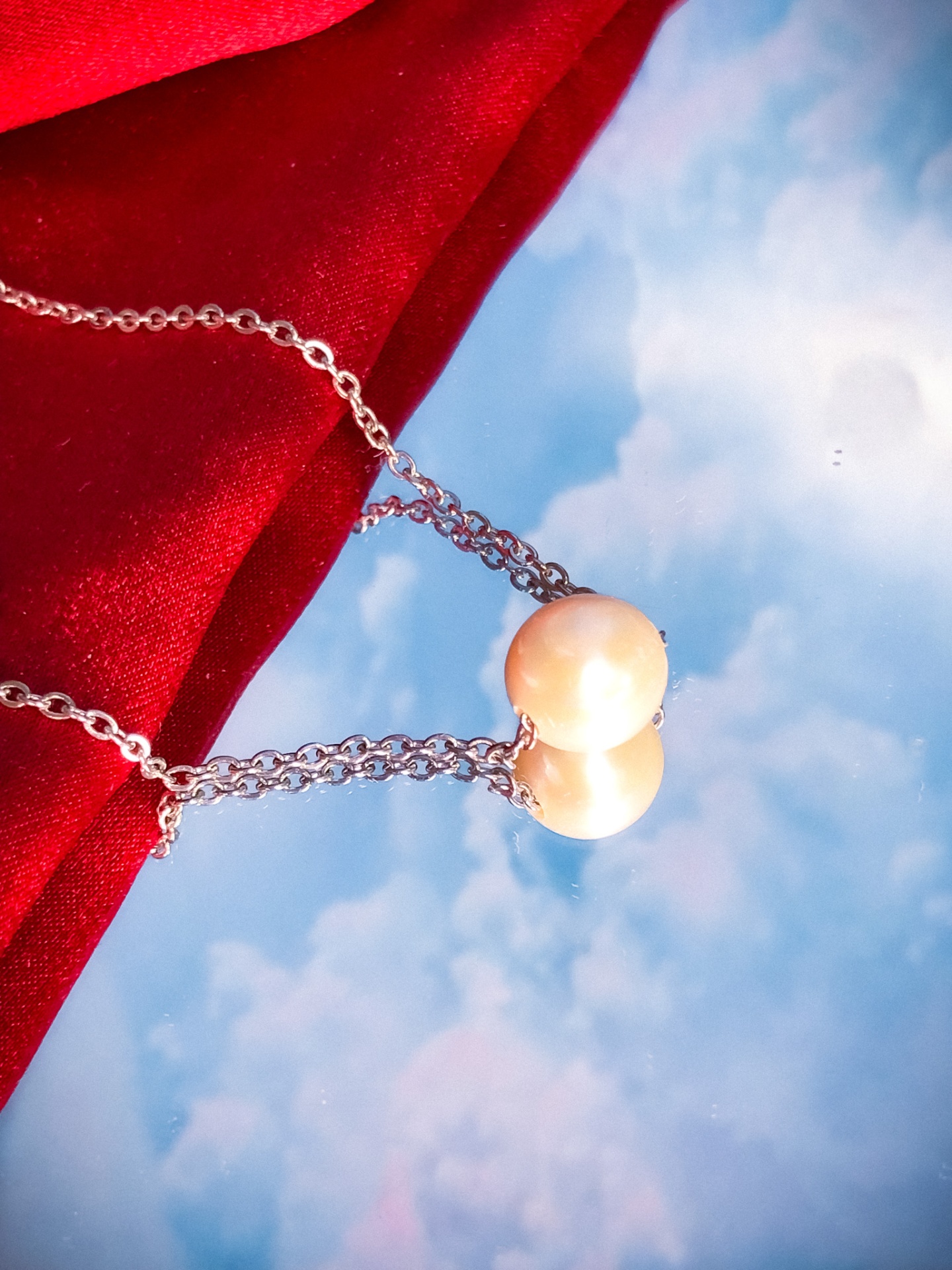 Colar com pérola branca e corrente prateada sobre reflexo do céu e tecido vermelho