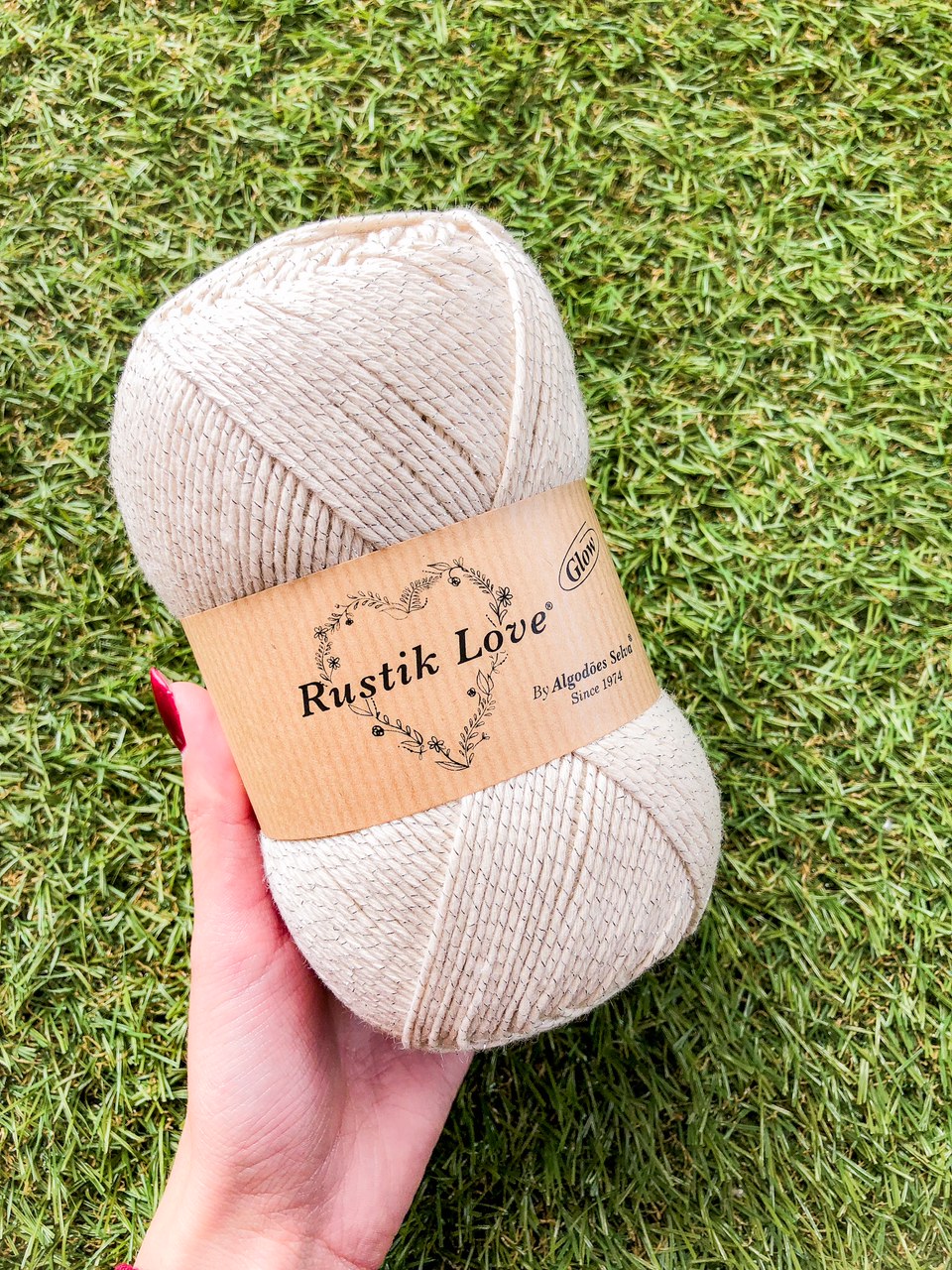 Novelo de fio bege claro com rótulo Rustik Love e fundo de relva verde