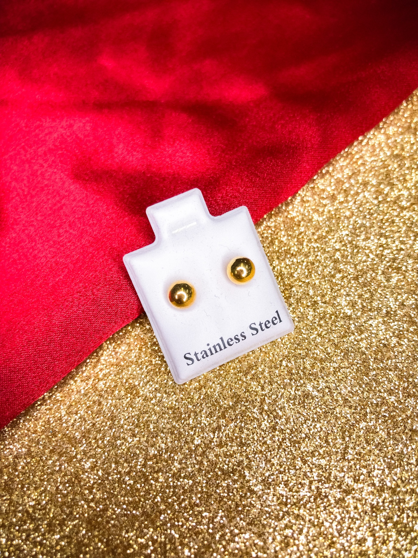 Brincos dourados redondos em suporte branco com texto, sobre fundo vermelho e dourado brilhante