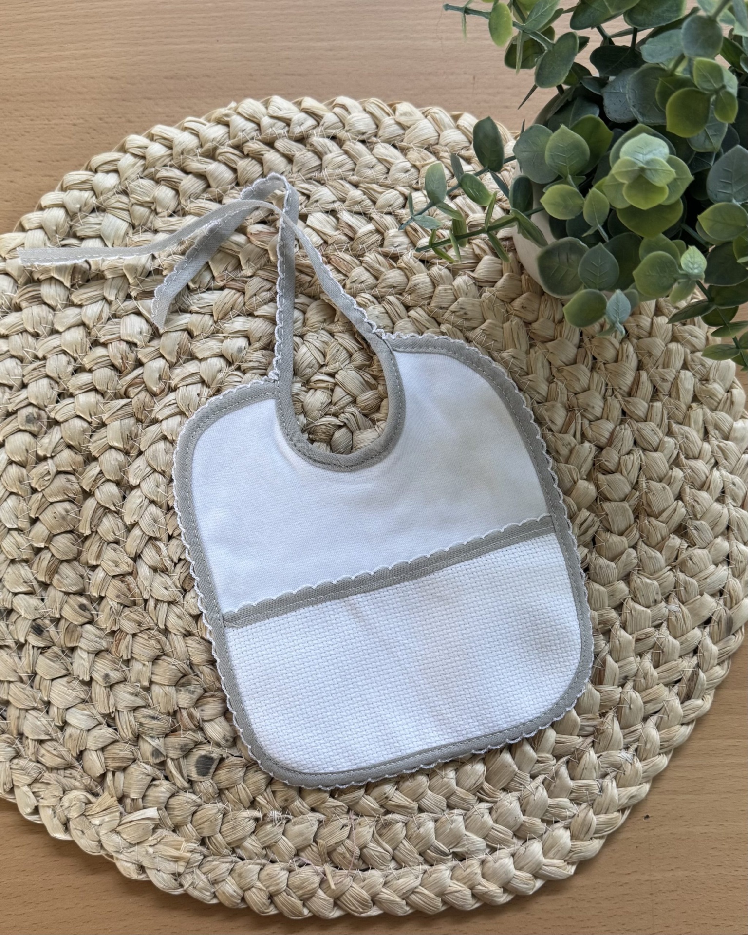 Babete branco com bordas cinzentas sobre tapete redondo de palha trançada com folhas verdes ao lado