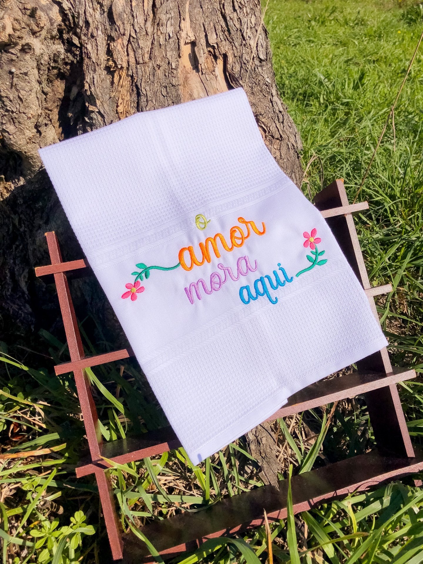 Toalha branca com bordado colorido 'amor mora aqui' em estrutura de ferro junto a tronco e relva