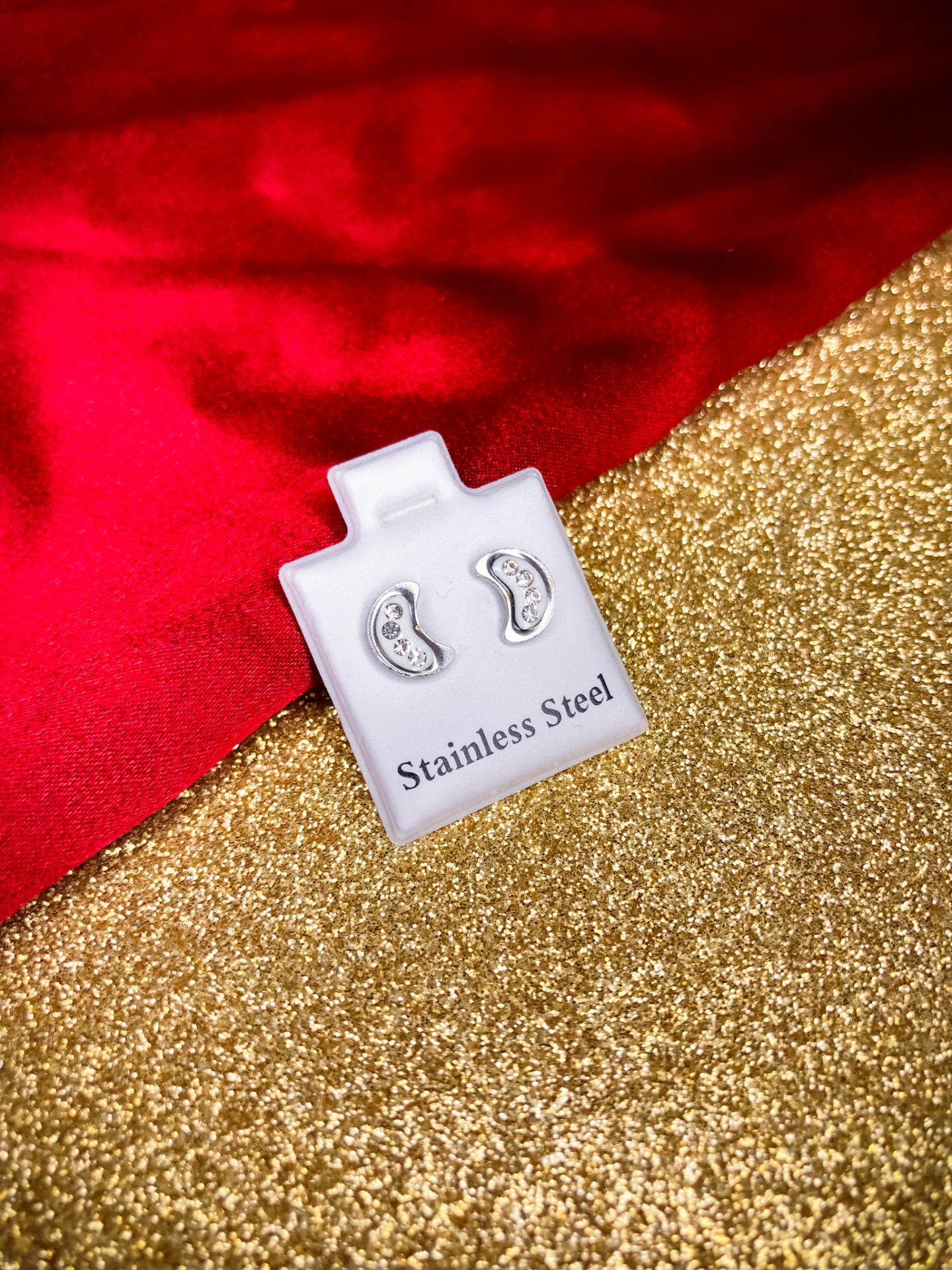 Brincos em aço inoxidável em suporte branco sobre fundo dourado brilhante e tecido vermelho