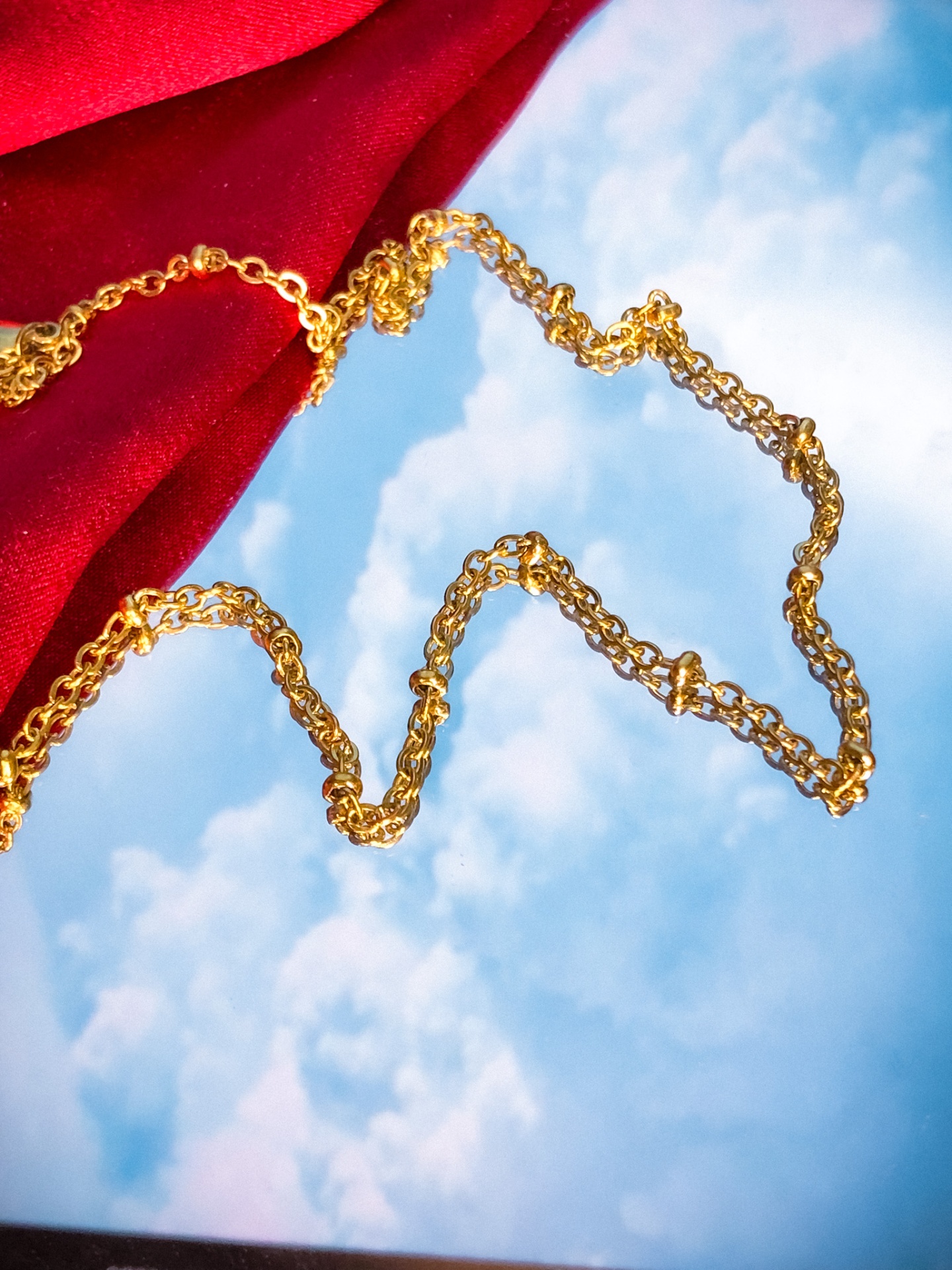 Colar dourado sobre fundo azul com nuvens e tecido vermelho