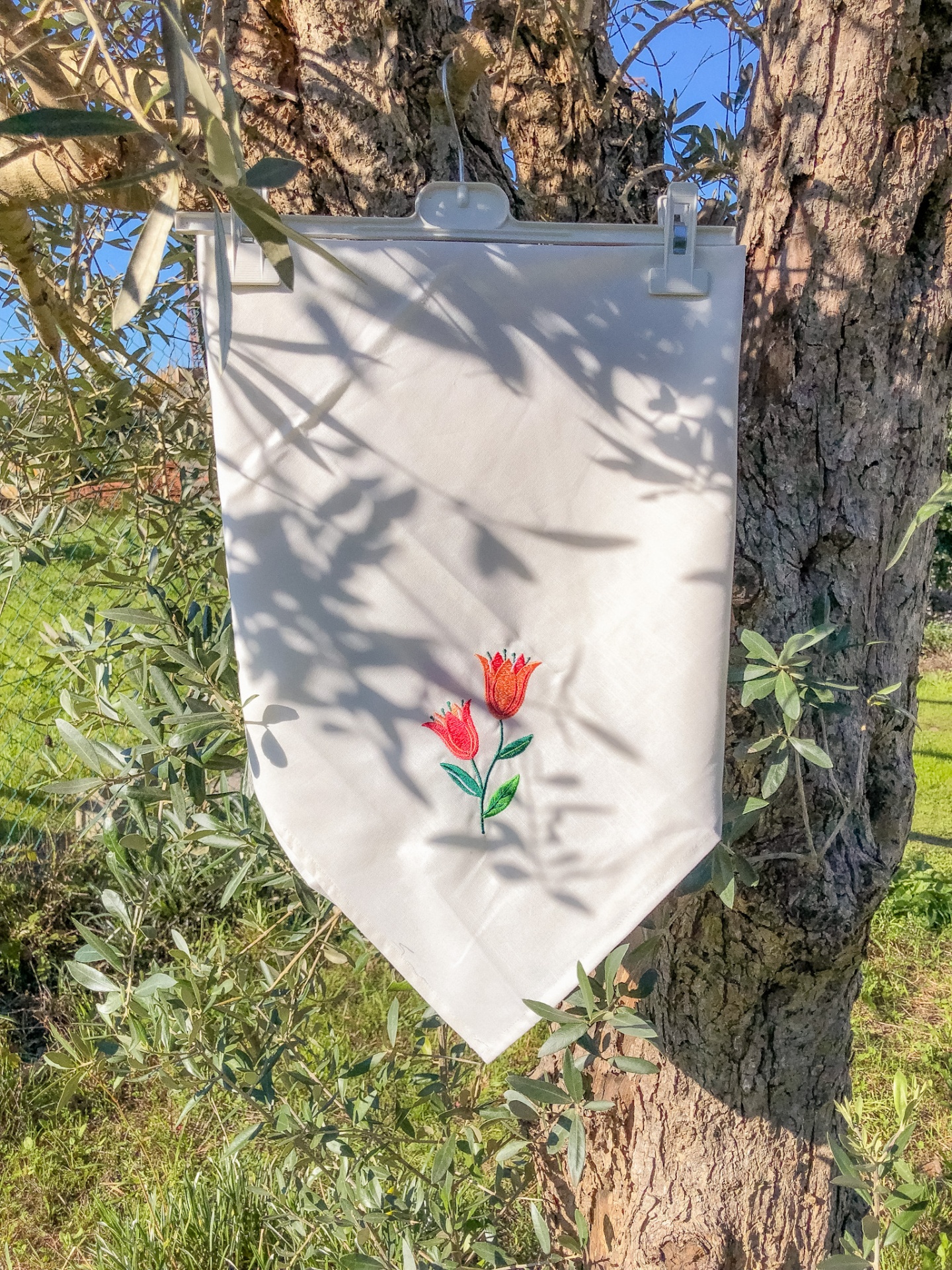 Bandeira branca com bordado de flores vermelhas pendurada numa árvore