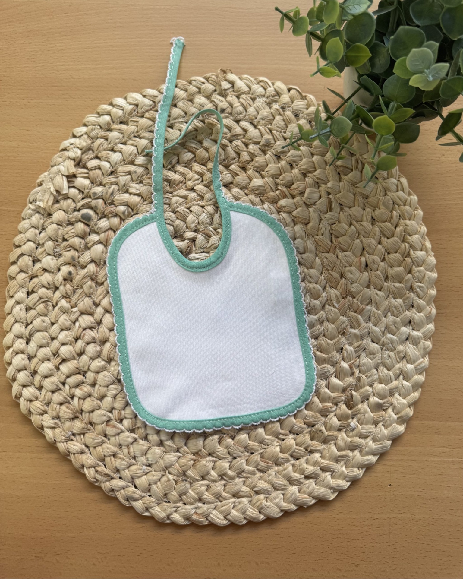 Babete branco com borda verde claro e fita para apertar sobre suporte trançado, planta verde ao lado