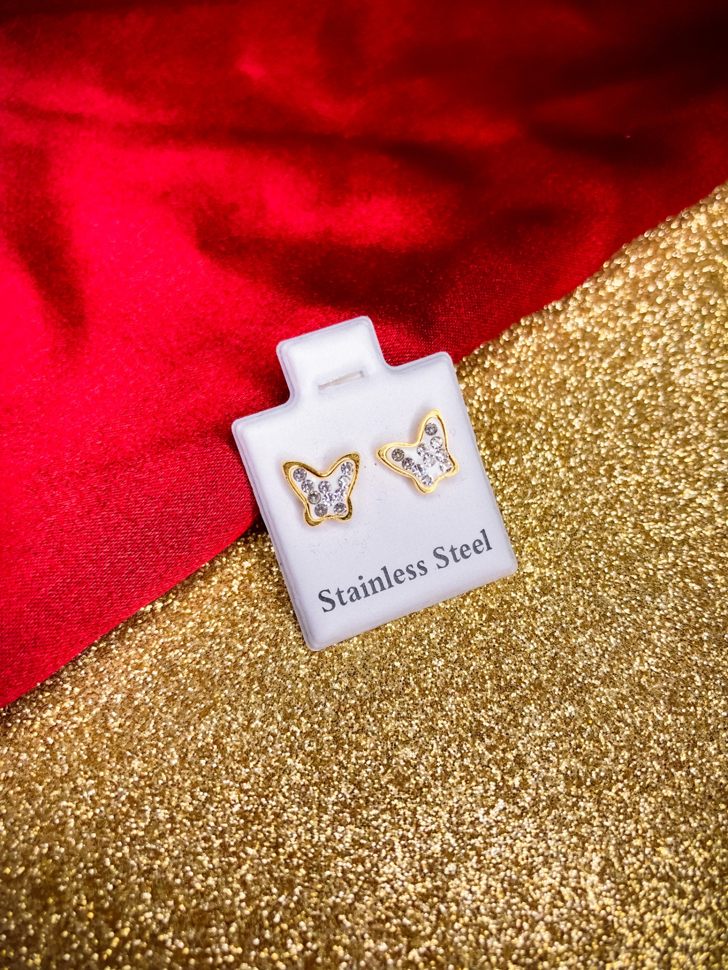 Brincos em aço inoxidável com design de borboleta e pedras brilhantes num suporte branco