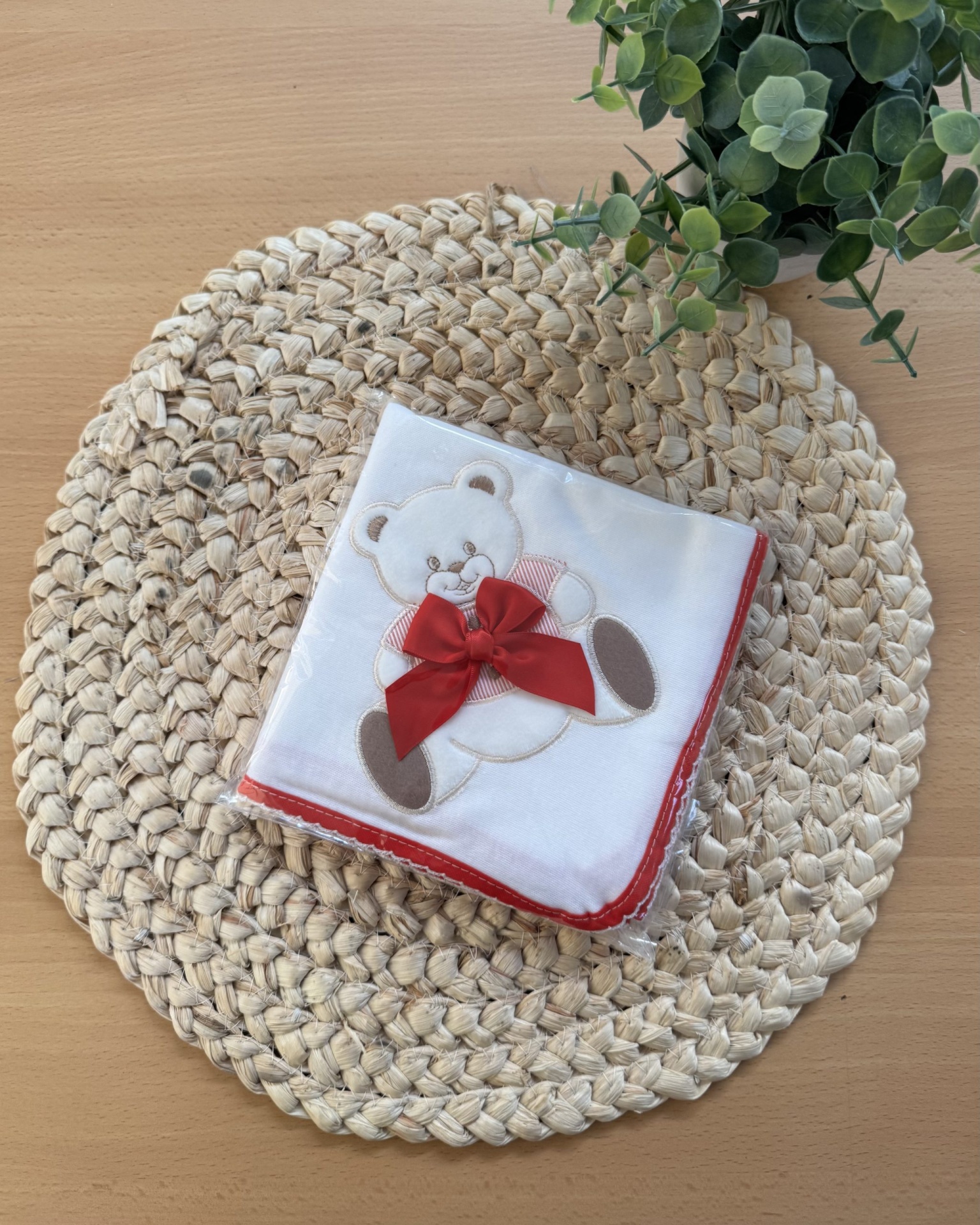 Fralda branca com ursinho bordado e laço vermelho sobre ráfia trançada e mesa de madeira com planta.