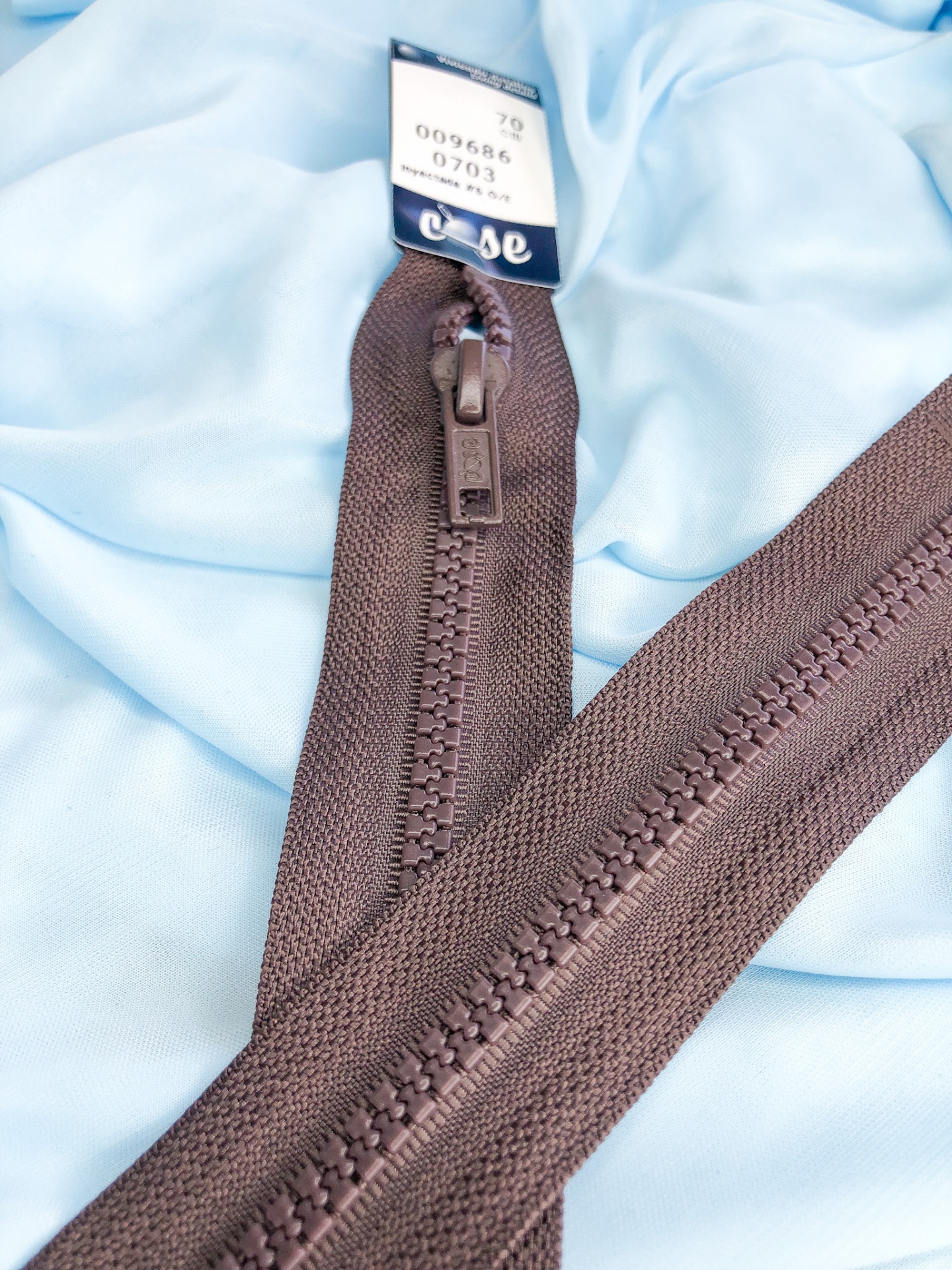 Zíper castanho escuro sobre tecido azul claro com etiqueta visível