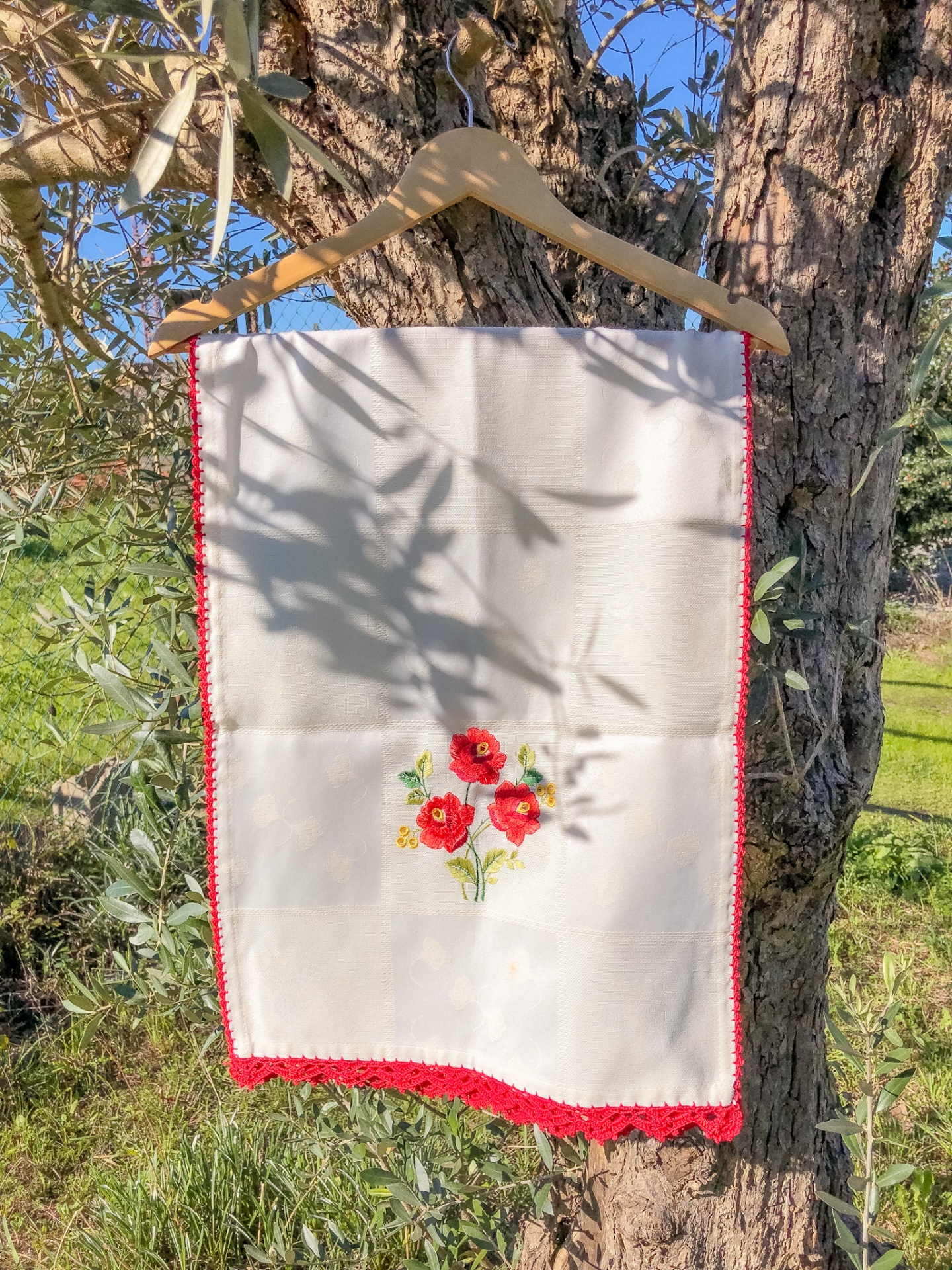 Toalha branca bordada com flores vermelhas pendurada num cabide de madeira numa árvore