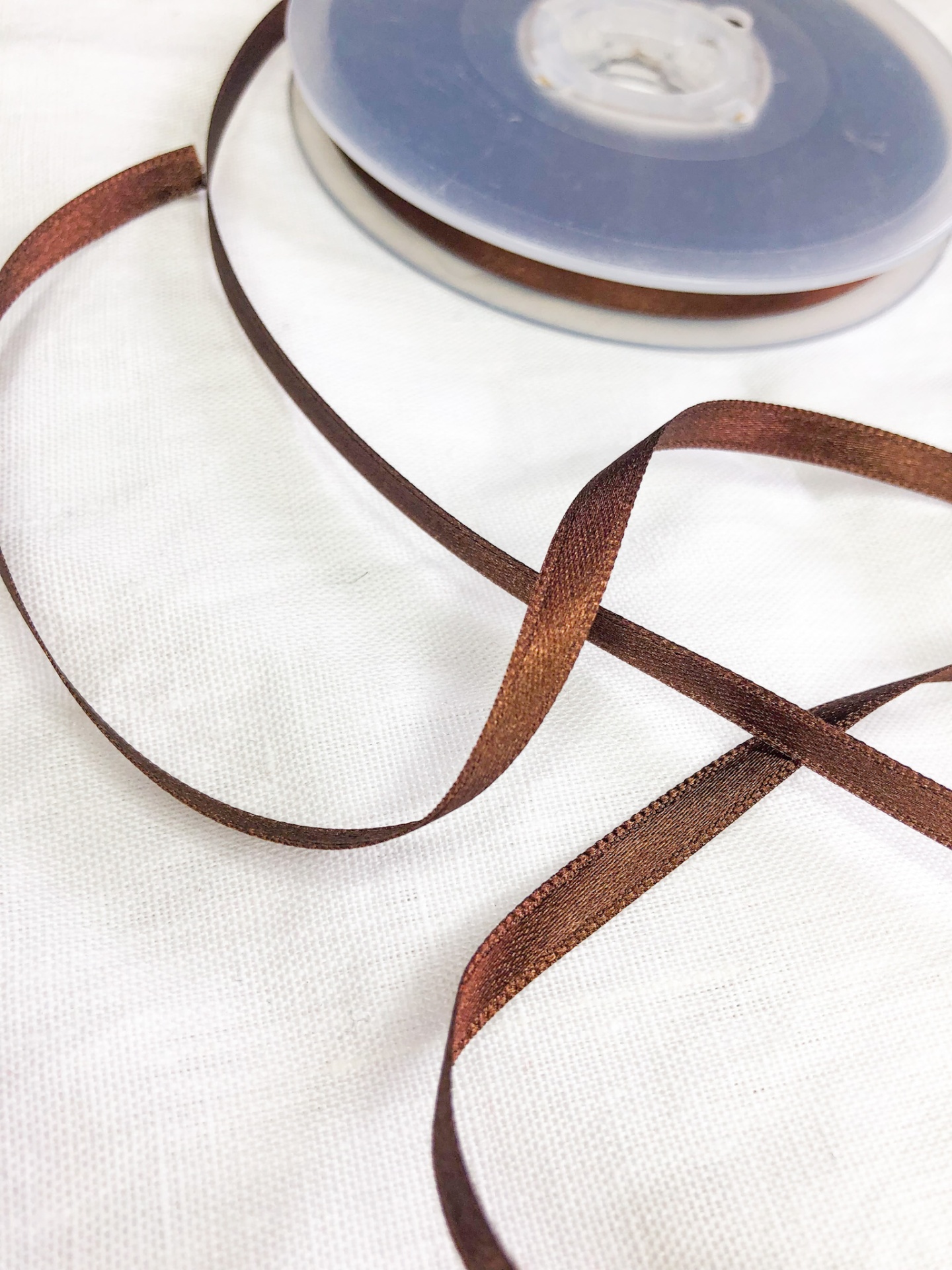 Fita de cetim castanha enrolada num rolo de plástico sobre tecido branco.