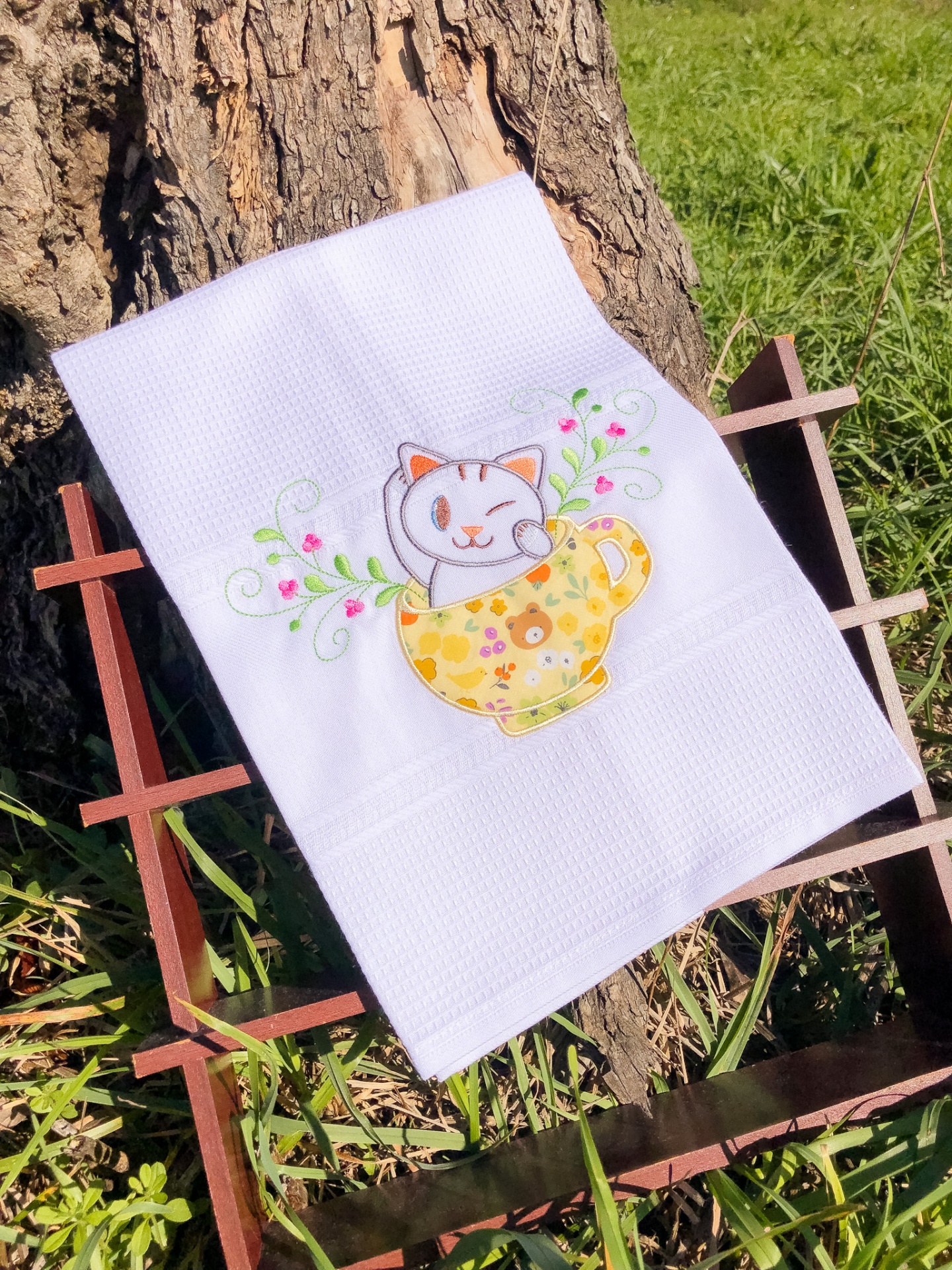 Toalha branca bordada com gato numa chávena amarela, em exposição ao ar livre num suporte de madeira.