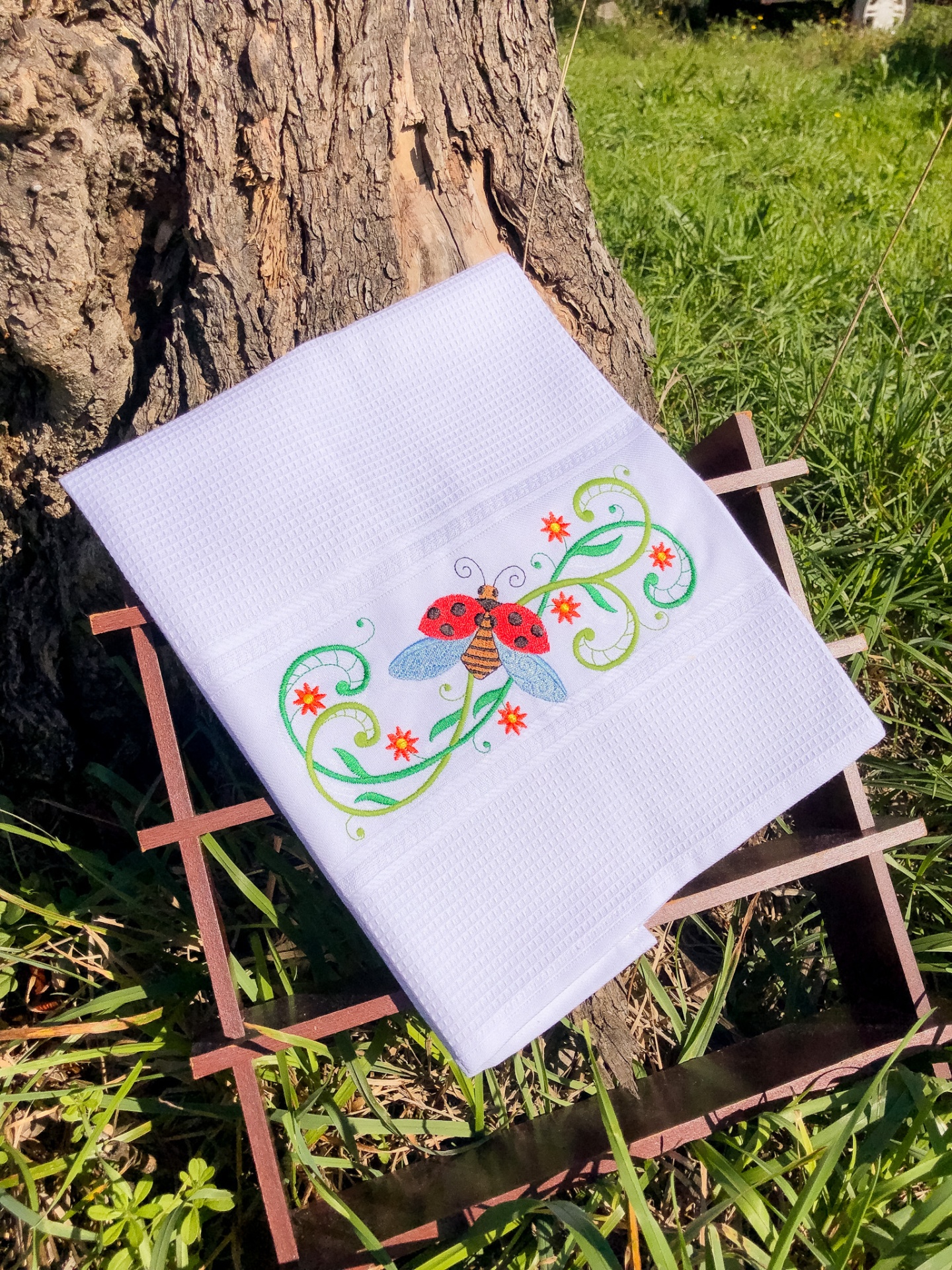 Toalha branca com bordado de joaninha, folhas e flores sobre estrutura de madeira, ao ar livre