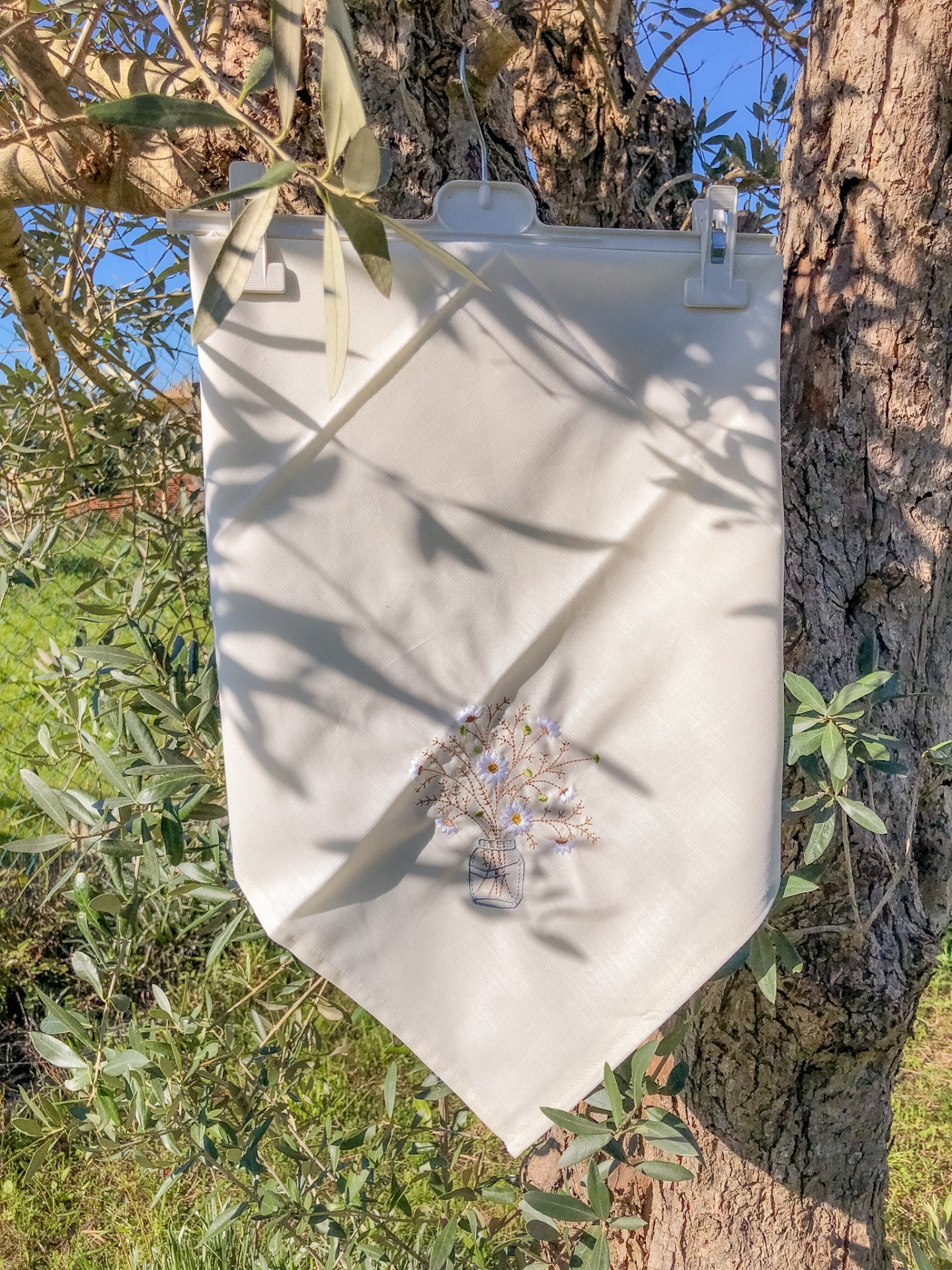 Pano decorativo branco com bordado de jarro com flores pendurado numa árvore