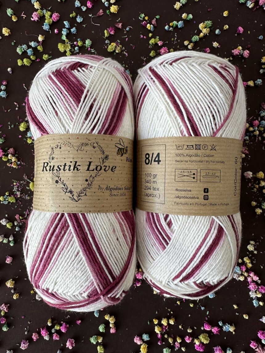 Dois novelos de fio branco e bordô com rótulo Rustik Love de algodão, sobre fundo escuro com flores pequenas coloridas.