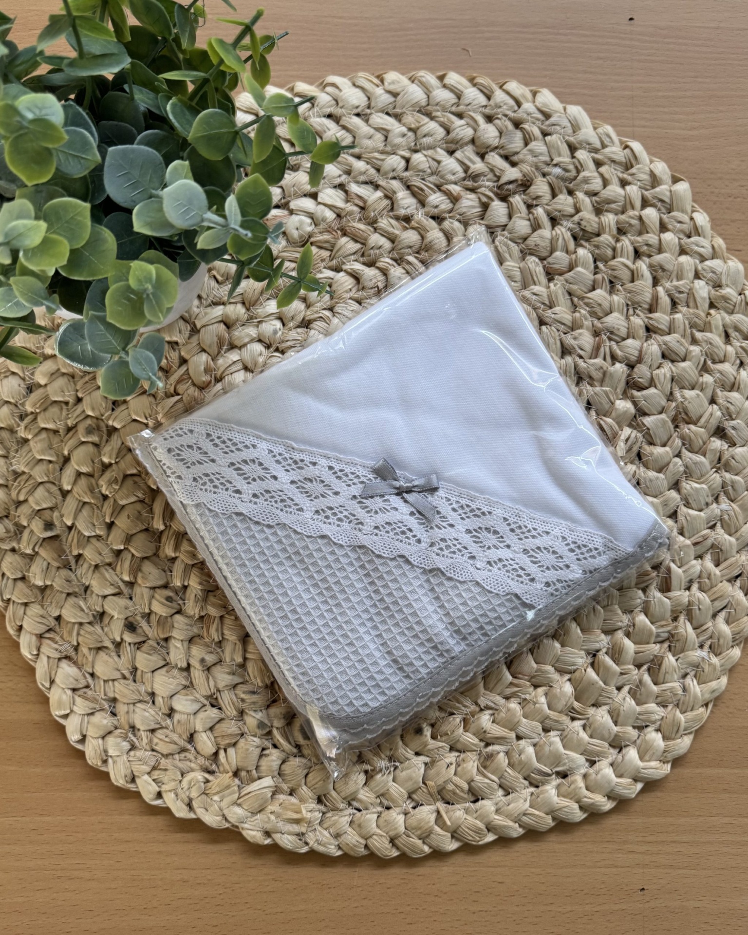 Toalha branca com borda cinzenta, renda e laço, em embalagem transparente sobre tapete de fibras naturais com planta