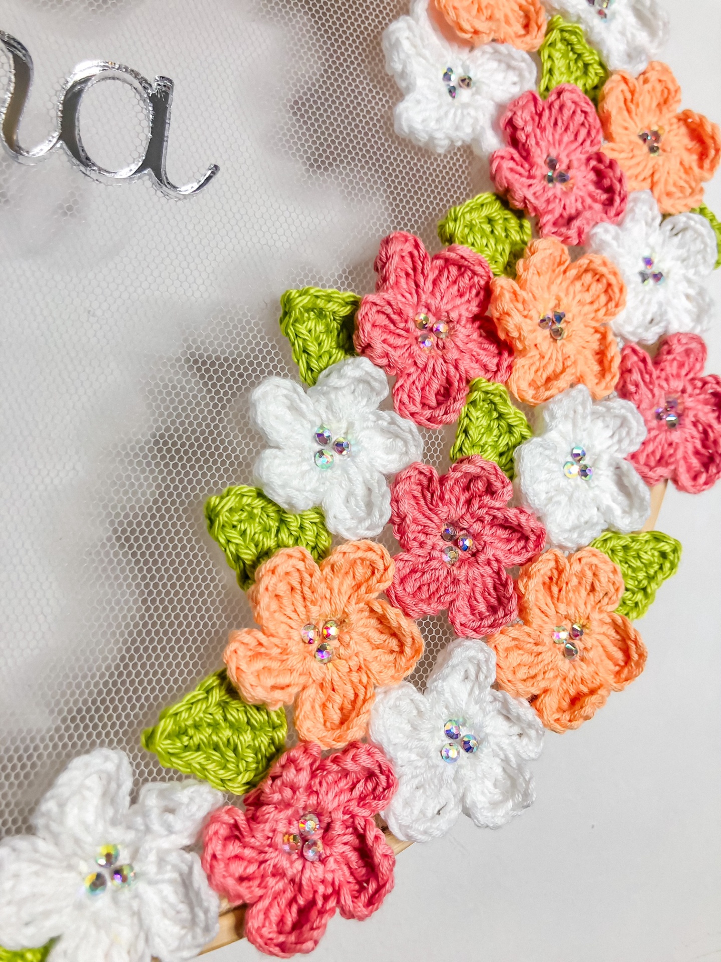 Guirlanda de croché com flores coloridas e folhas verdes, com miçangas no centro das flores.