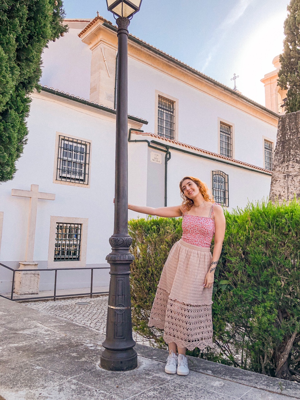 Mulher com vestido floral vermelho e saia bege em exterior junto a poste e edifício branco.