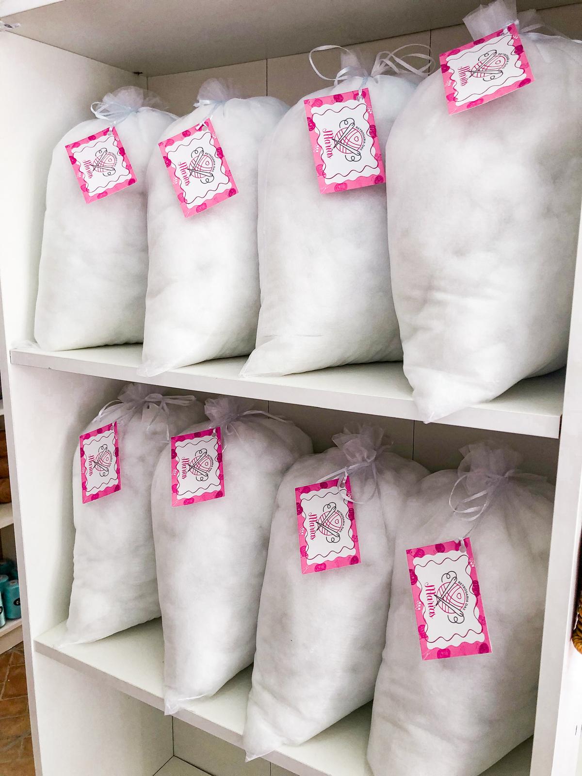 Sacos brancos com enchimento e etiquetas rosa pendurados em prateleiras brancas.