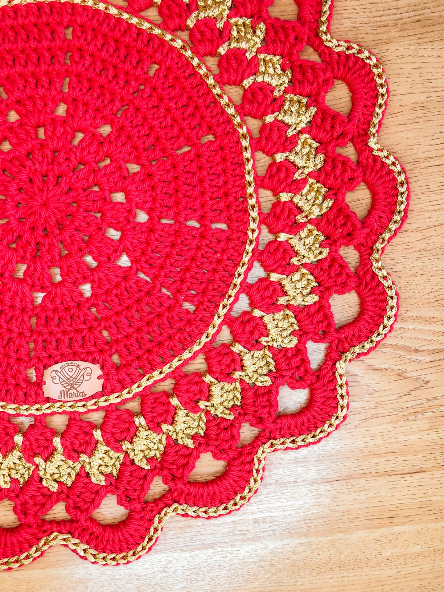 Toalha redonda de croché vermelha e dourada com etiqueta Maine sobre mesa de madeira clara