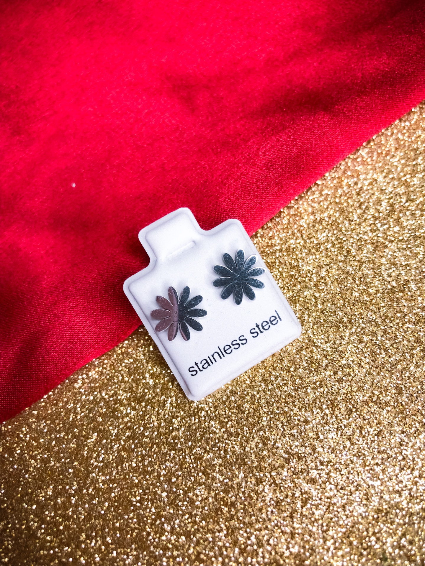 Brincos de flor em aço inoxidável sobre cartão branco com texto, fundo vermelho e dourado glitter