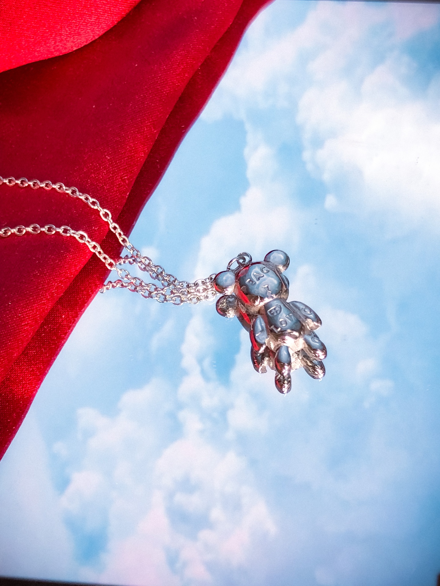 Colar prateado com pendente de urso sobre tecido vermelho e fundo de céu com nuvens