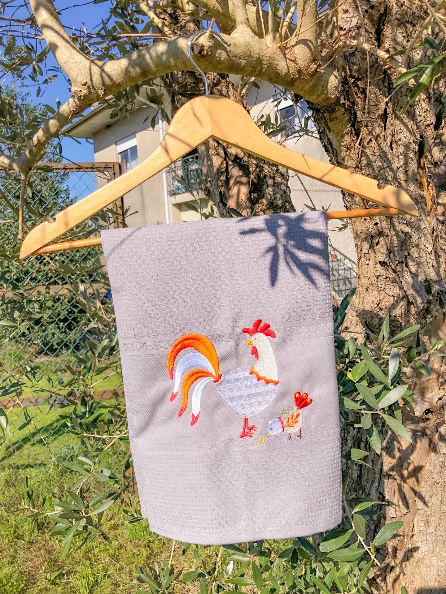 Toalha cinzenta com bordado de galo pendurada em cabide numa árvore
