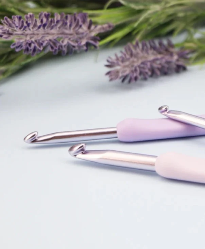 Duas agulhas de croché roxas com flores de lavanda ao fundo