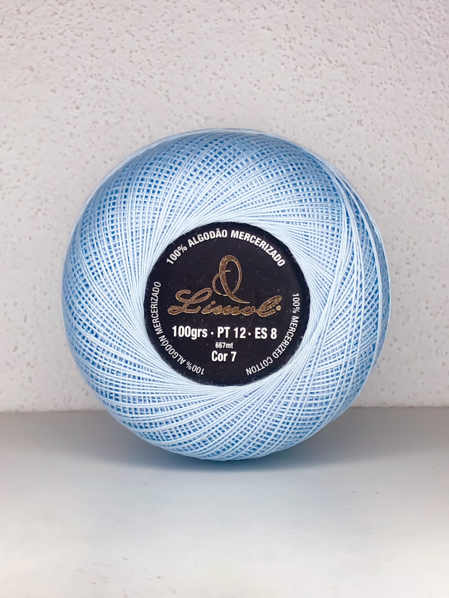 Bola de linha azul clara com etiqueta preta com texto sobre superfície branca e parede bege