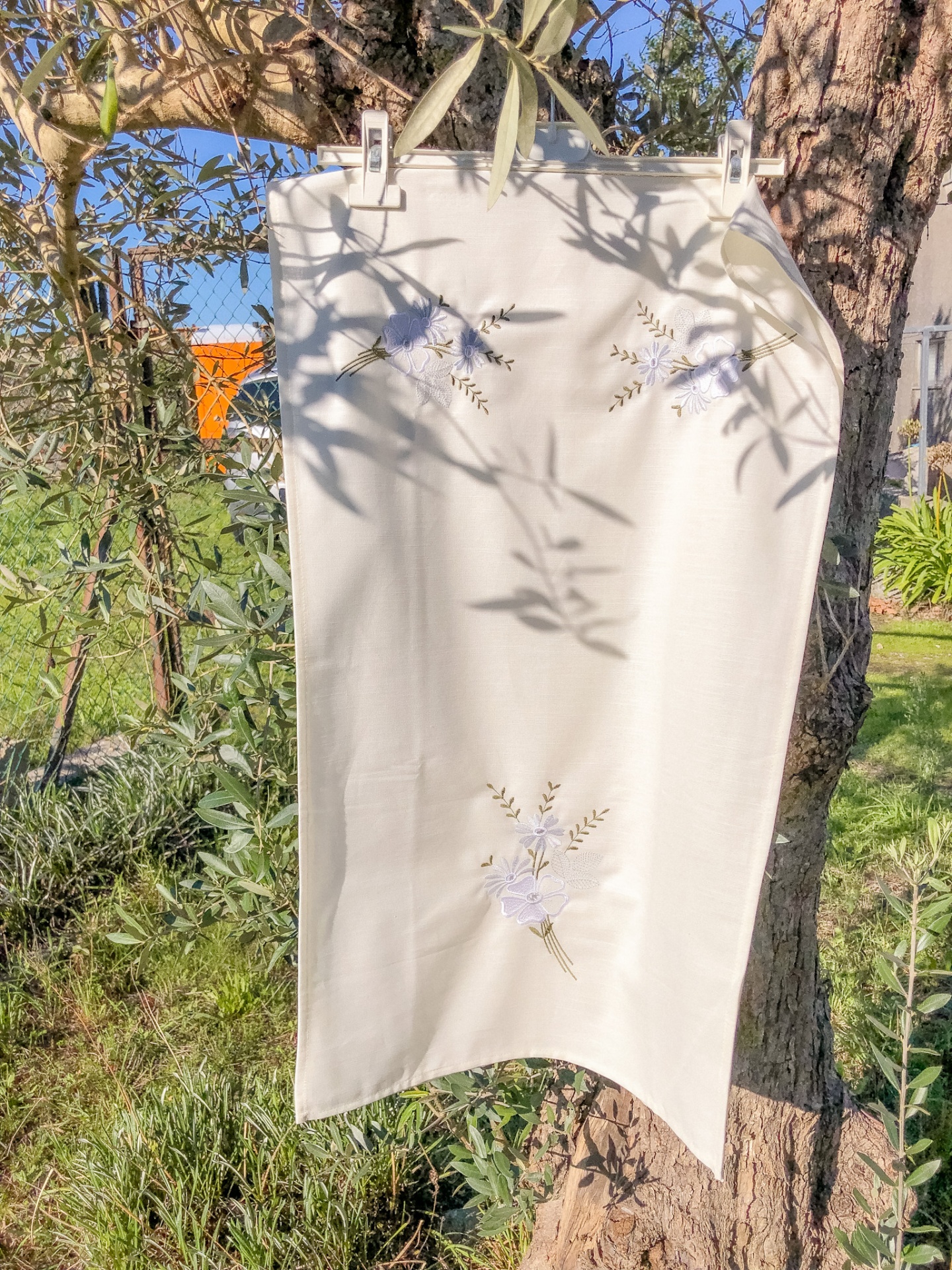 Toalha branca com bordados florais pendurada ao sol junto a árvore