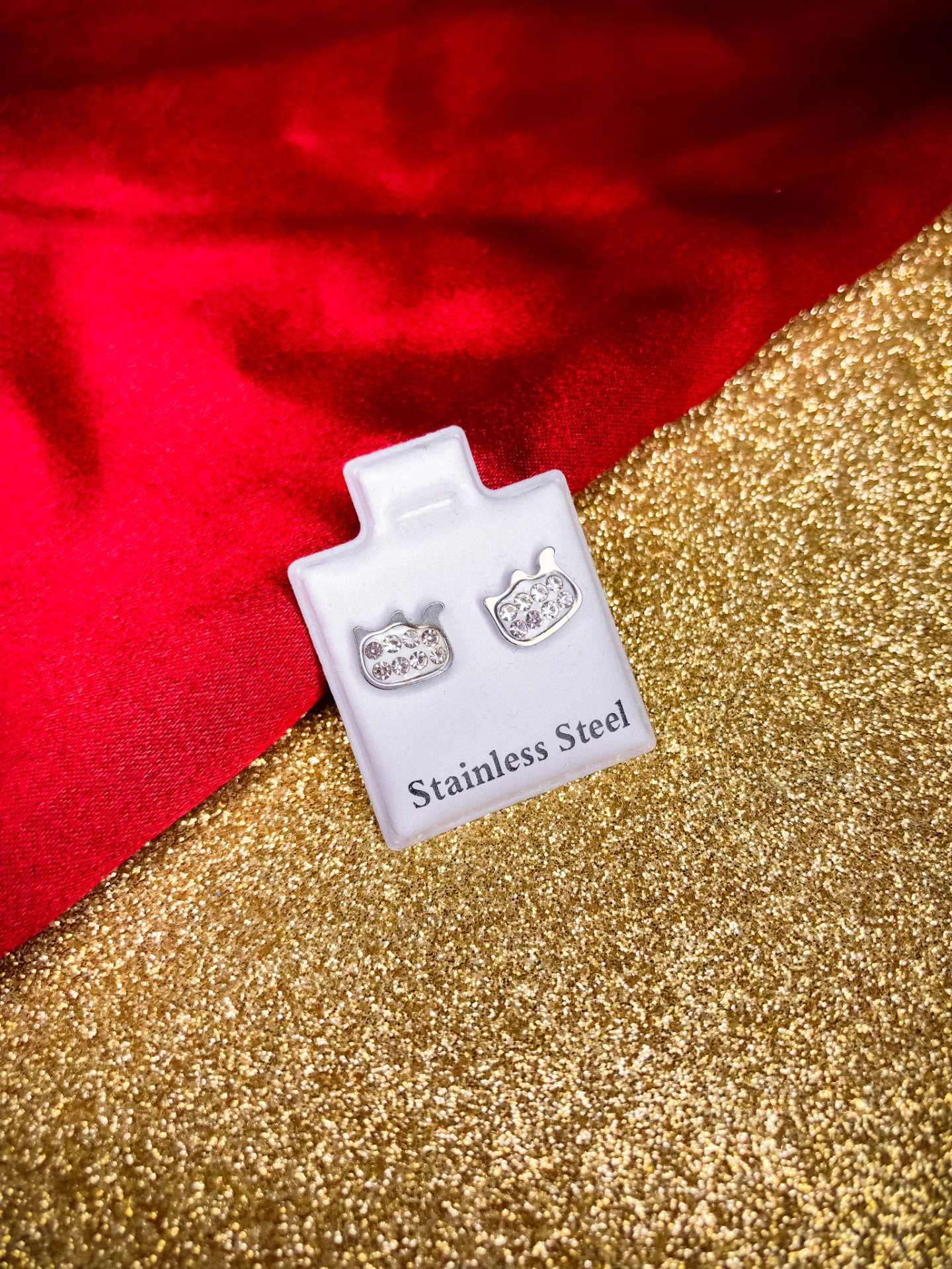 Brincos em aço inoxidável com forma de abóbora com pedras brilhantes em base branca com texto sobre fundo dourado e tecido vermelho