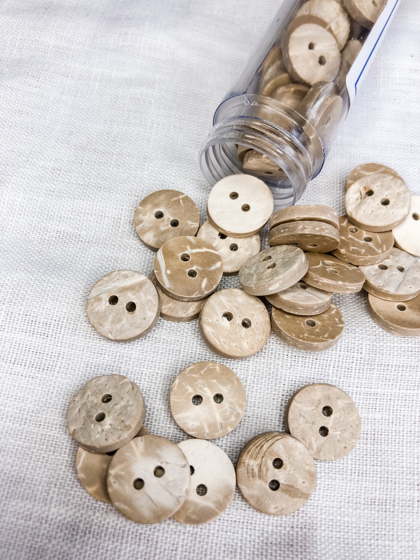 Botões de madeira clara com dois buracos sobre tecido branco e em recipiente plástico
