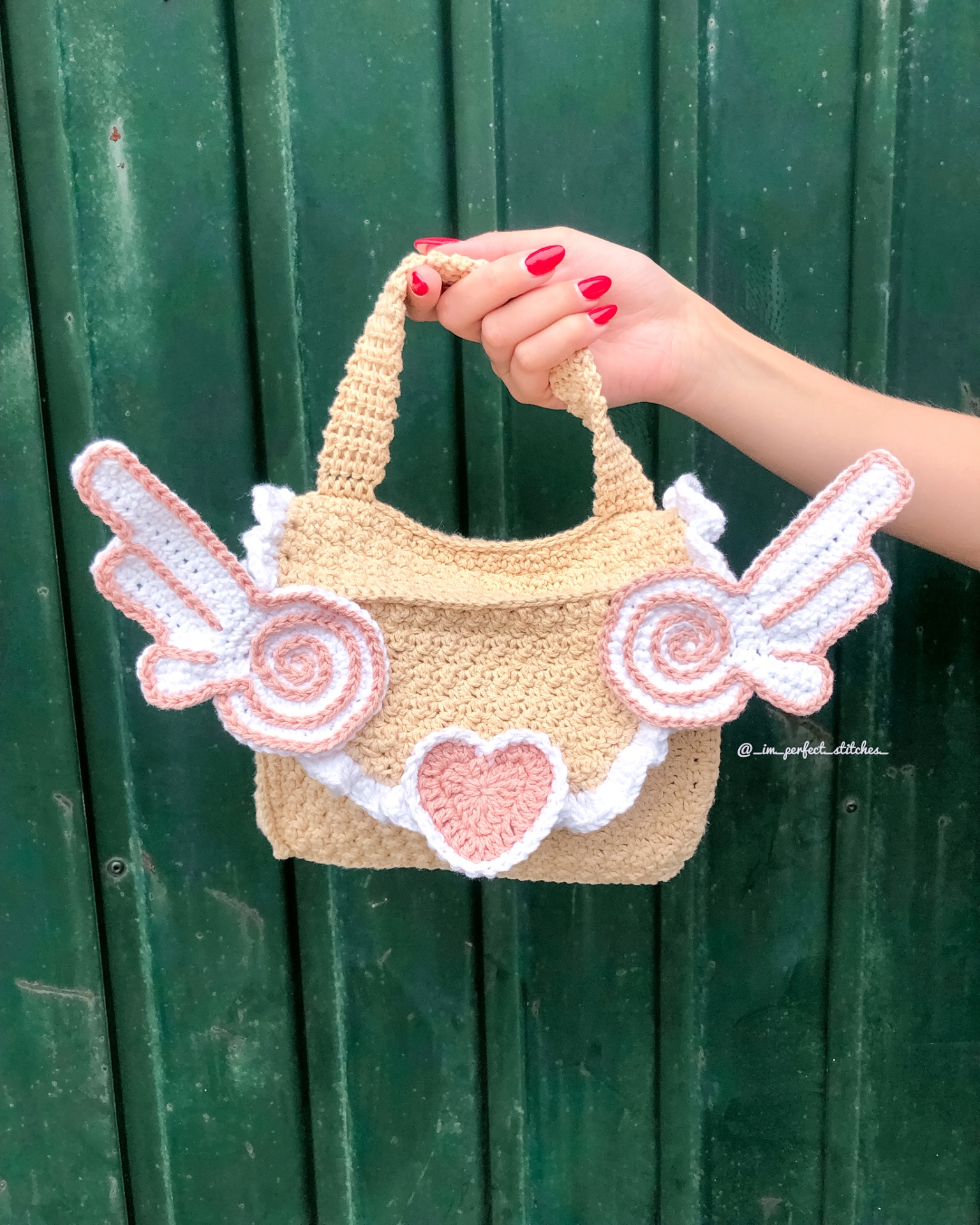 Mala de mão em crochet bege com asas e coração em rosa e branco contra porta verde.
