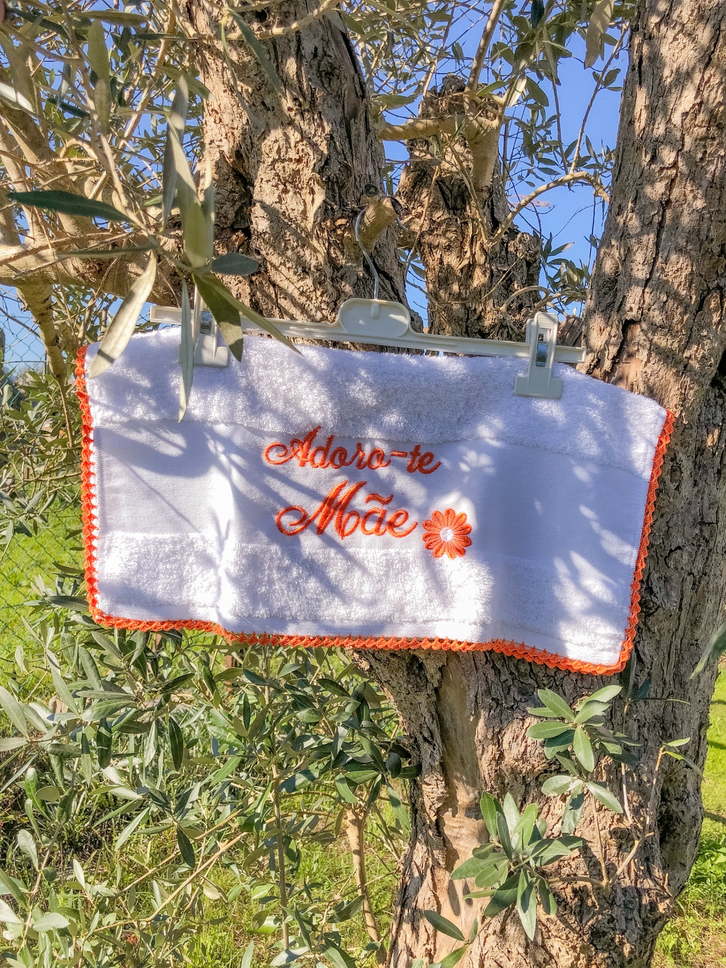 Toalha branca bordada a laranja com texto 'Adoro-te Mãe' pendurada num tronco de árvore