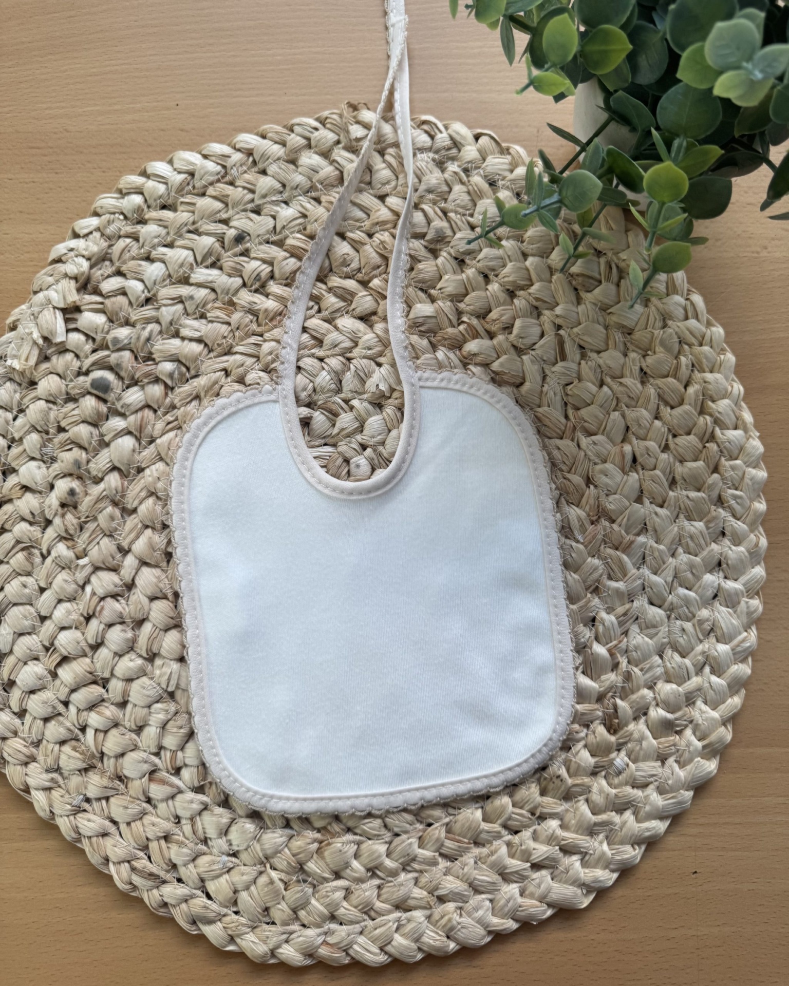 Babete branco com rebordo bege em apoio trançado redondo e plantas ao lado