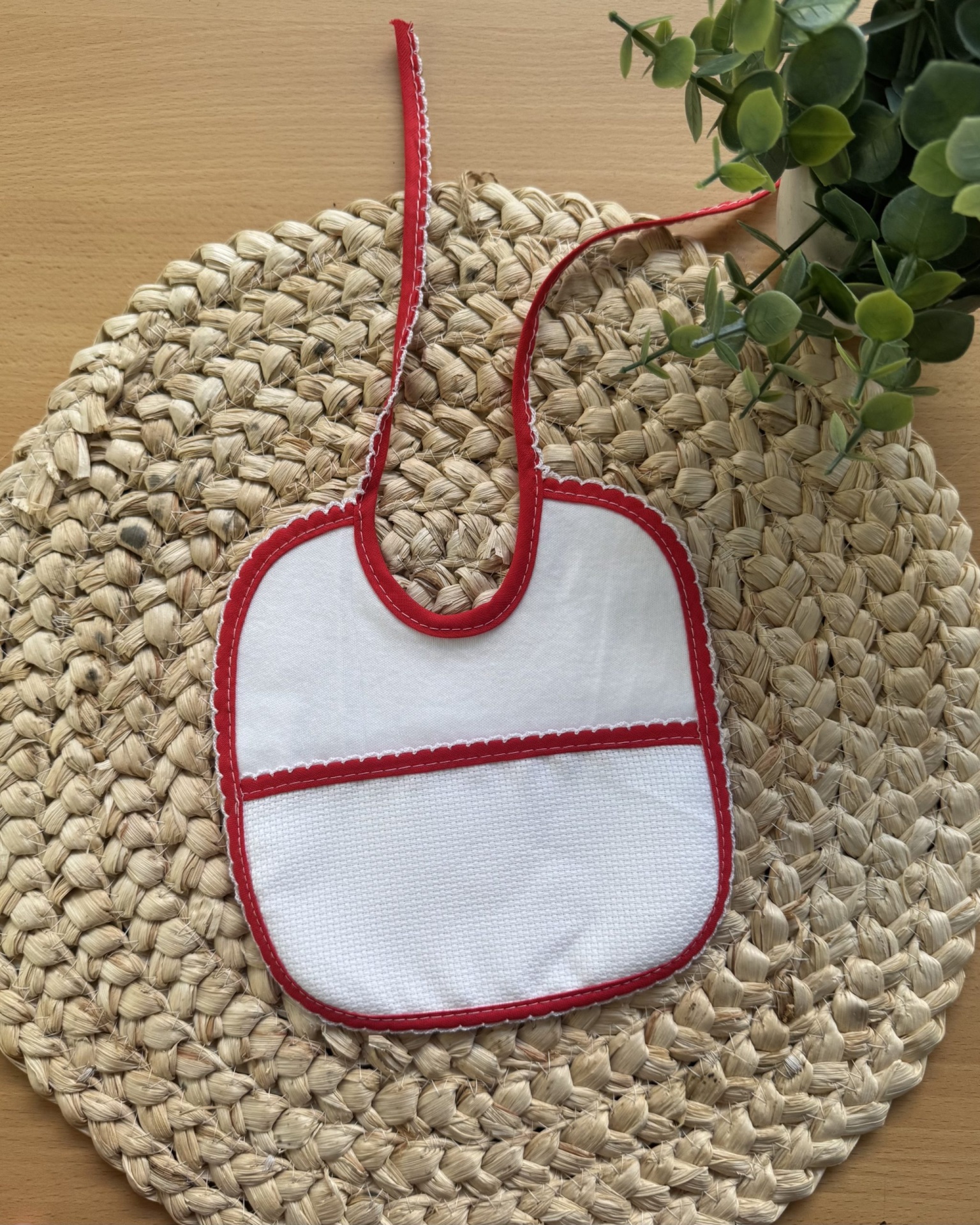 Babete branco com bordas vermelhas sobre base de fibra natural e planta verde
