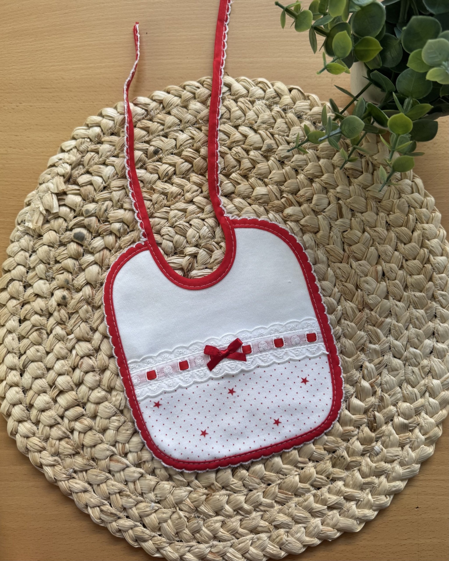 Babete infantil branco com bordas vermelhas e laço, sobre base de palha com planta verde