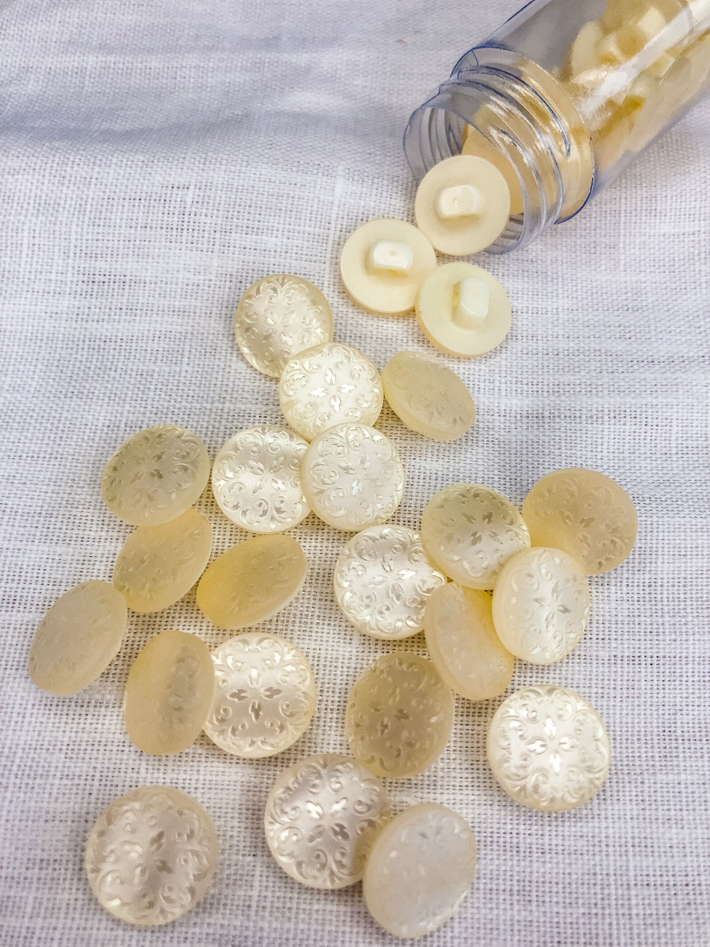 Botões redondos creme translúcidos com padrão floral sobre tecido branco, alguns dentro de tubo plástico