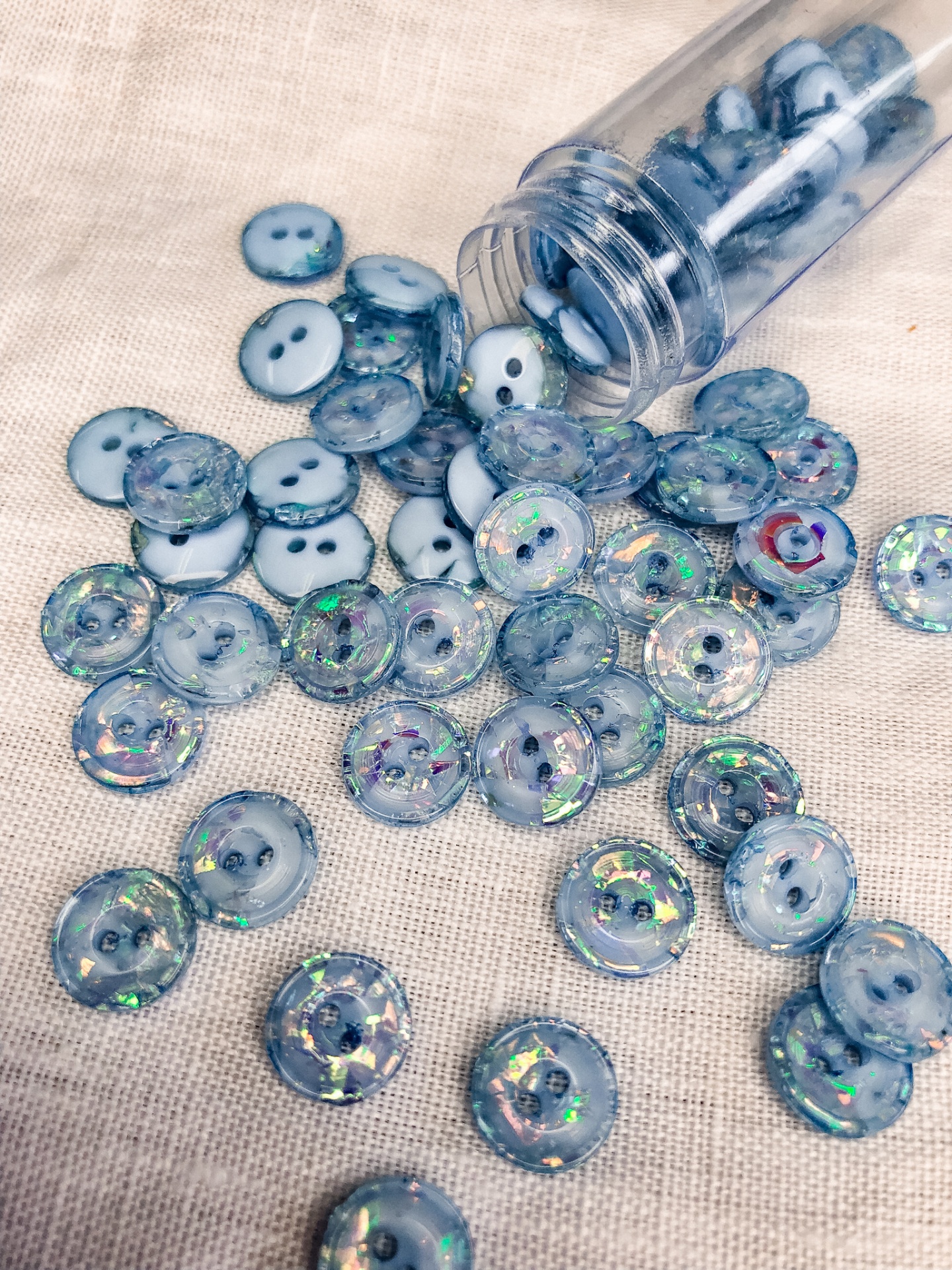 Botões azuis claros holográficos e opacos num tecido bege, com frasco plástico transparente