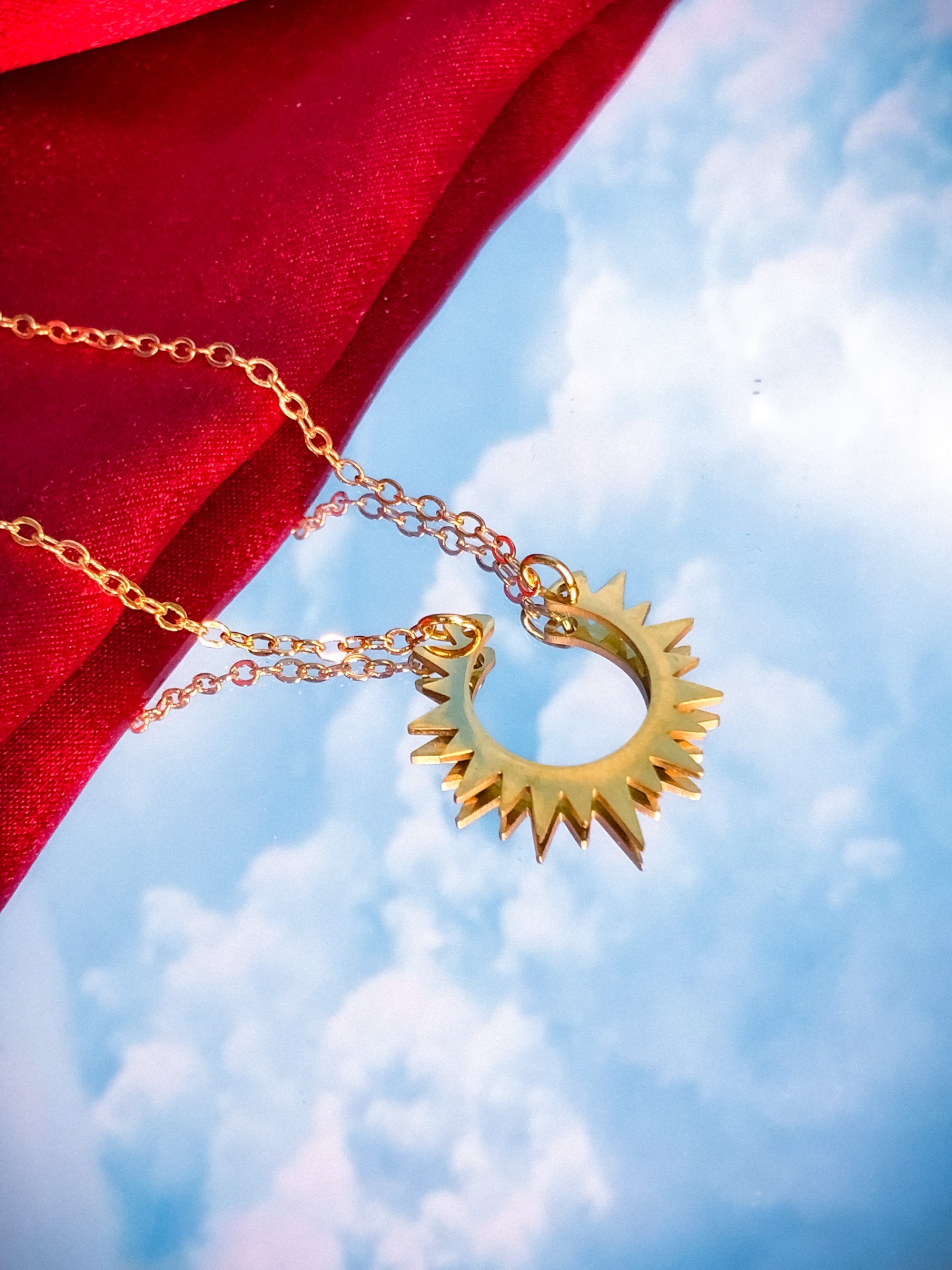 Colar dourado com pendente em forma de sol sobre fundo vermelho e céu azul