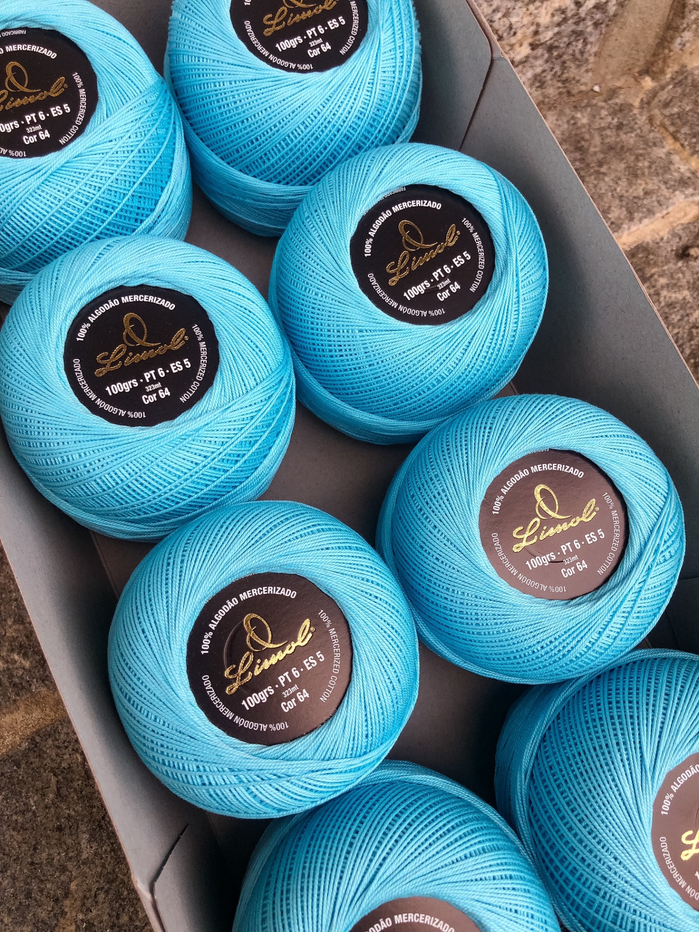 Oito novelos de linha azul claro com etiquetas pretas numa caixa cinzenta sobre pedra