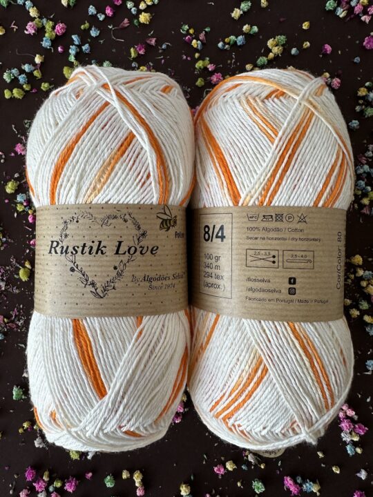 Dois novelos de fio de algodão branco e laranja com etiquetas Rustik Love sobre fundo escuro com flores coloridas.