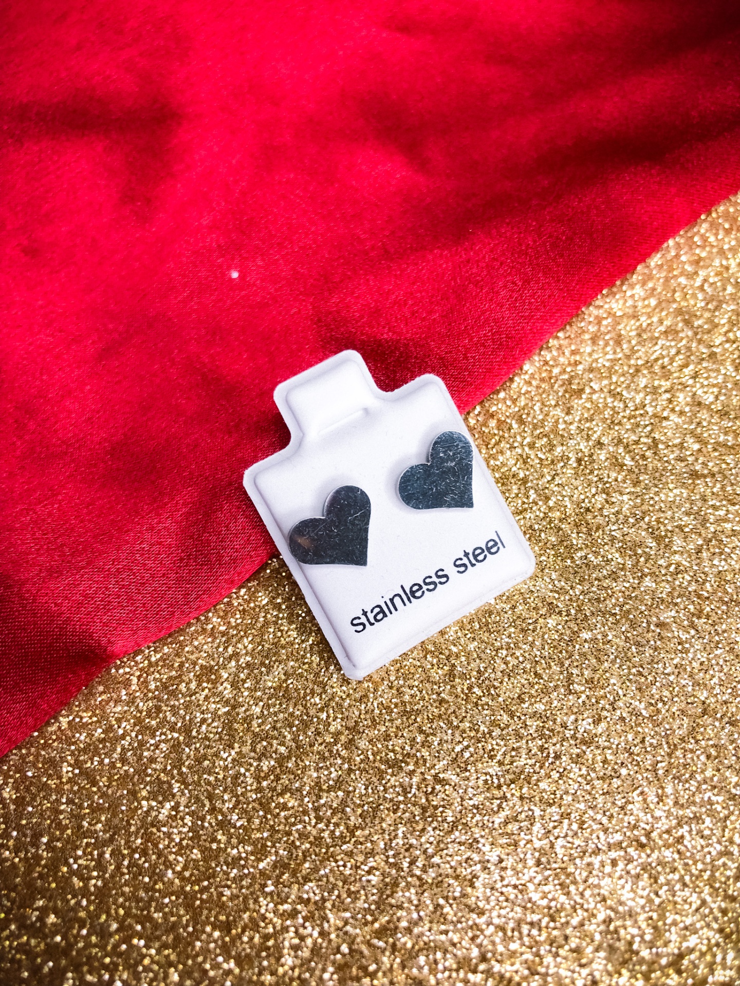 Brincos de aço inoxidável em formato de coração preto sobre suporte branco com texto, fundo vermelho e dourado.