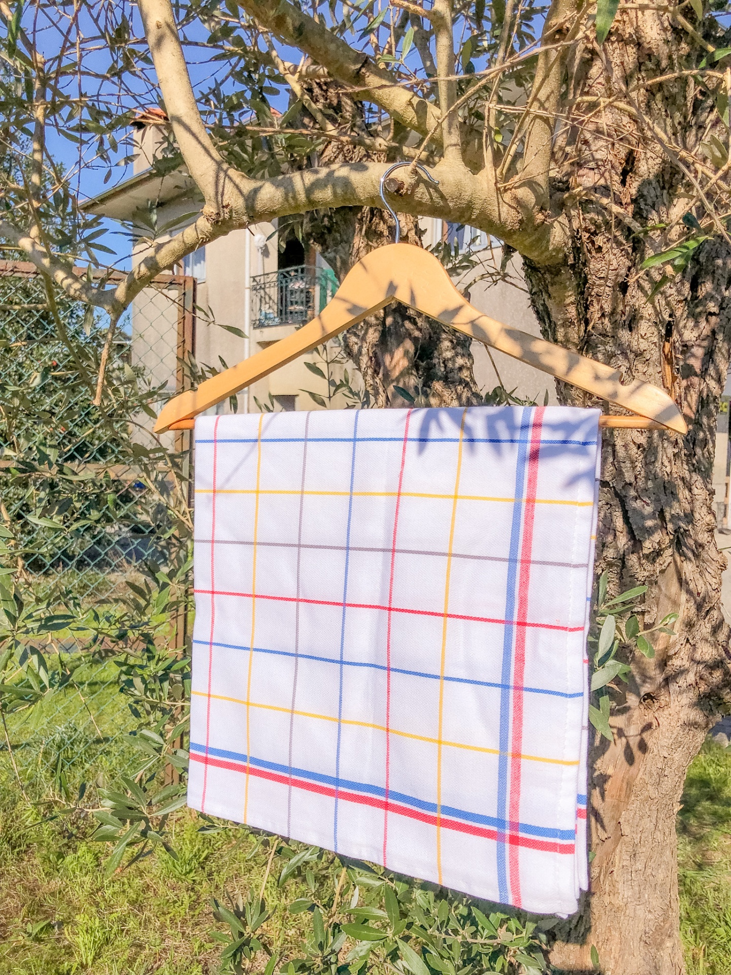 Pano de cozinha branco com linhas de grelha coloridas pendurado em cabide de madeira numa árvore ao ar livre
