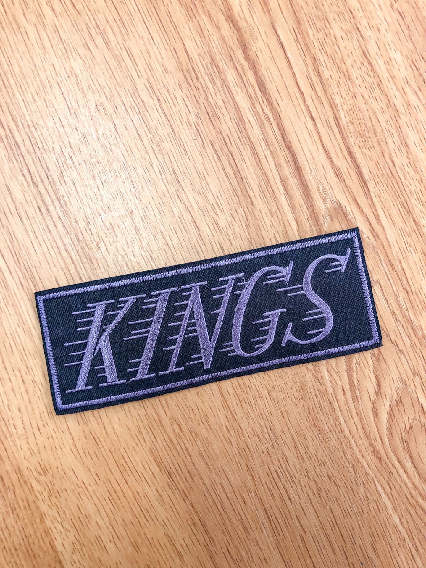 Patch de tecido preto com texto 'KINGS' cinzento sobre superfície de madeira clara
