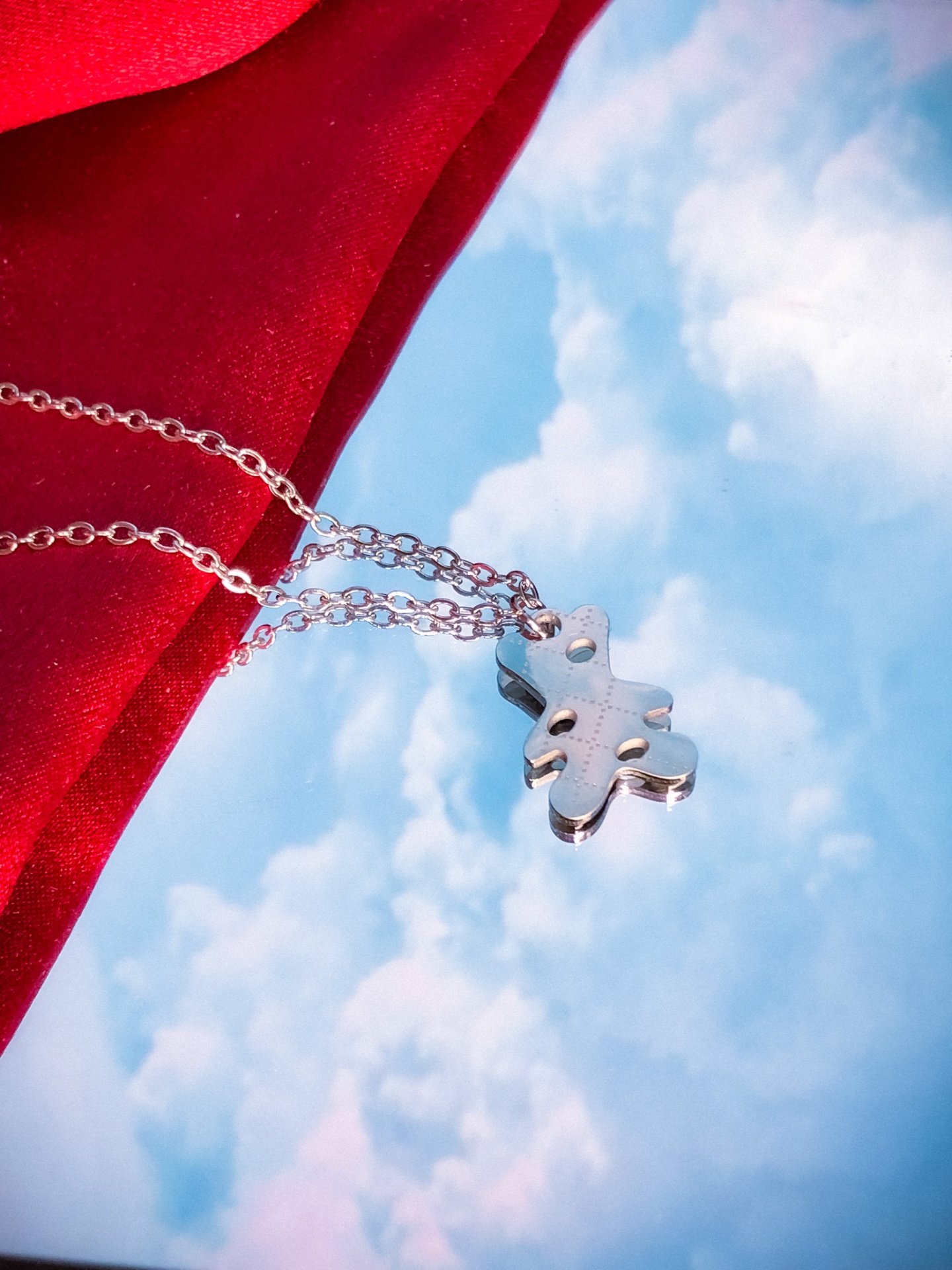 Colar prateado com pendente em forma de cruz contra céu azul com nuvens e tecido vermelho