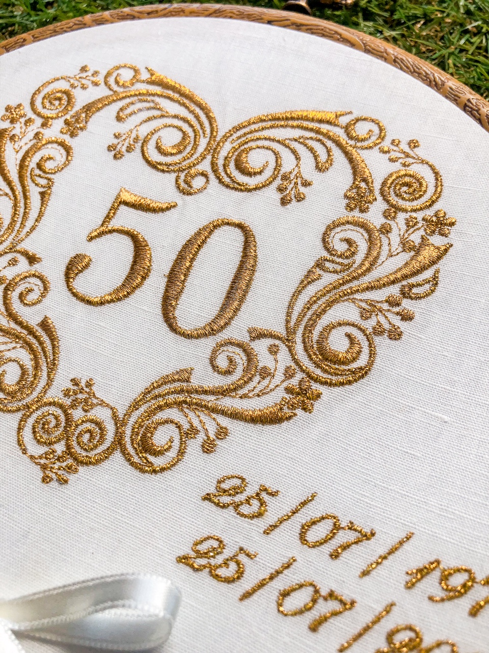 Bordado dourado sobre pano branco com número 50 e datas bordadas, em bastidor de madeira.